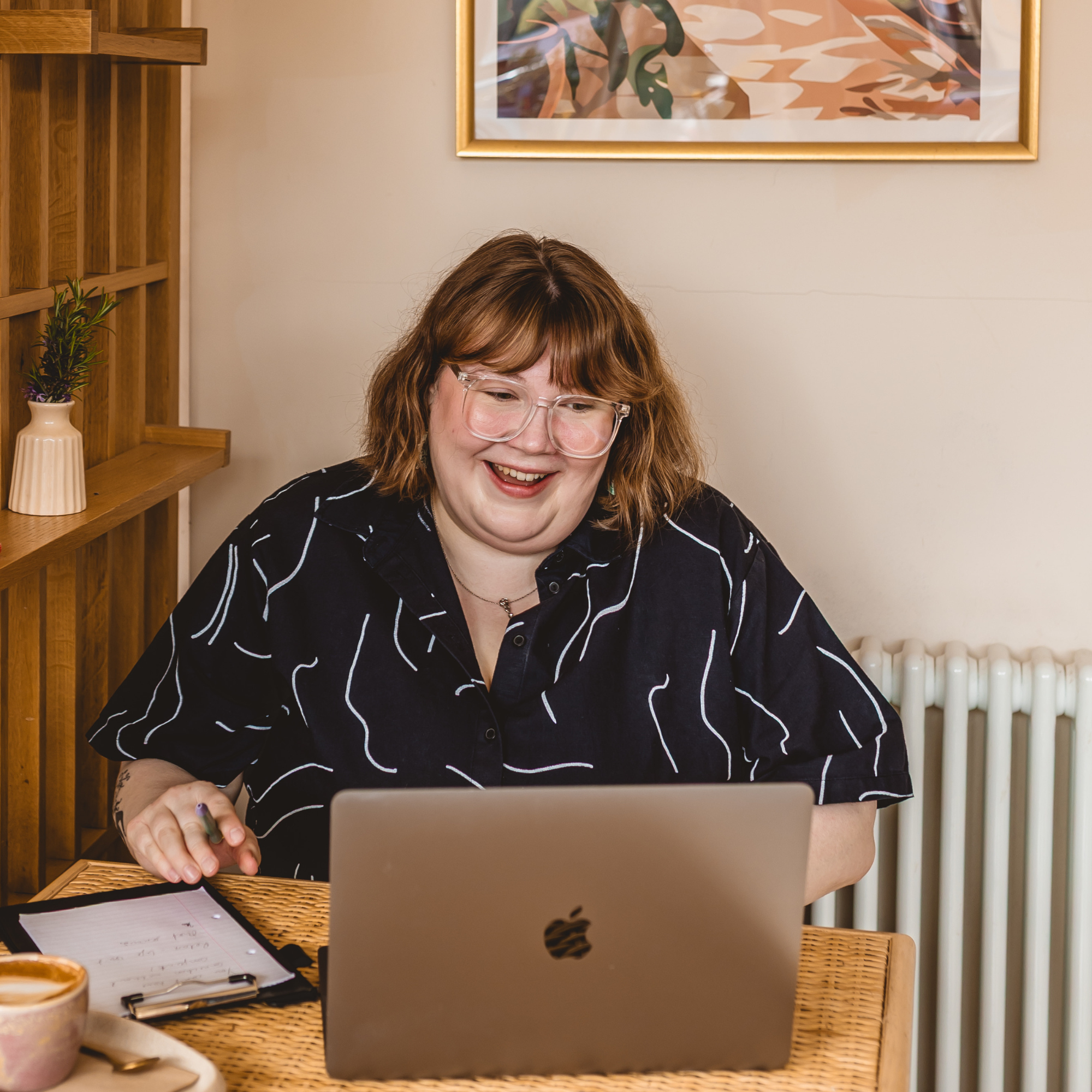 Woman with brown hair and glasses working on a laptop at a table, with a notebook, cup of coffee, and plant nearby.