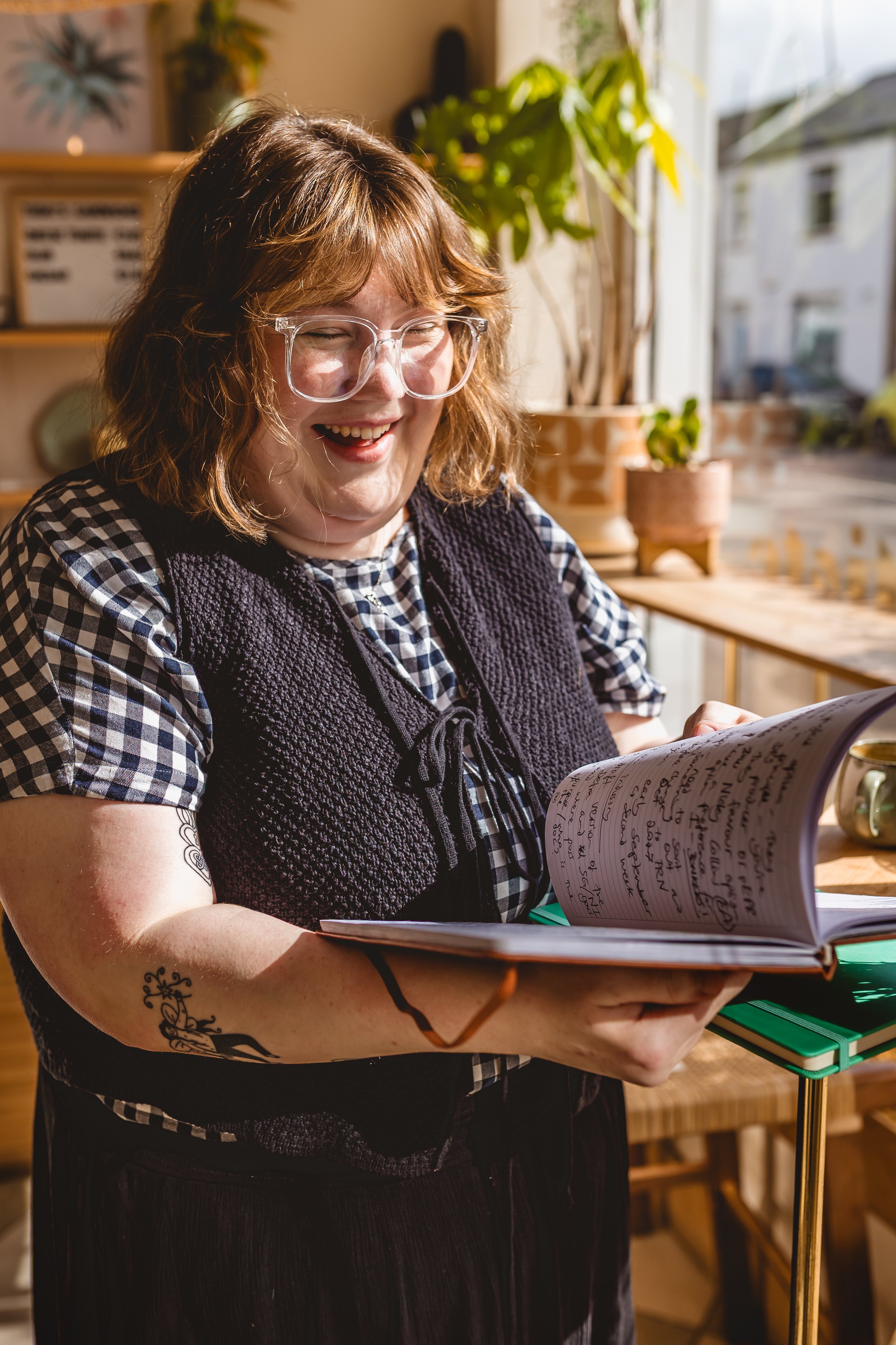 A woman with wavy, shoulder-length hair, glasses, and tattoos, smiling as she looks at a notebook full of handwritten notes, with sunlight streaming in through a window behind her.