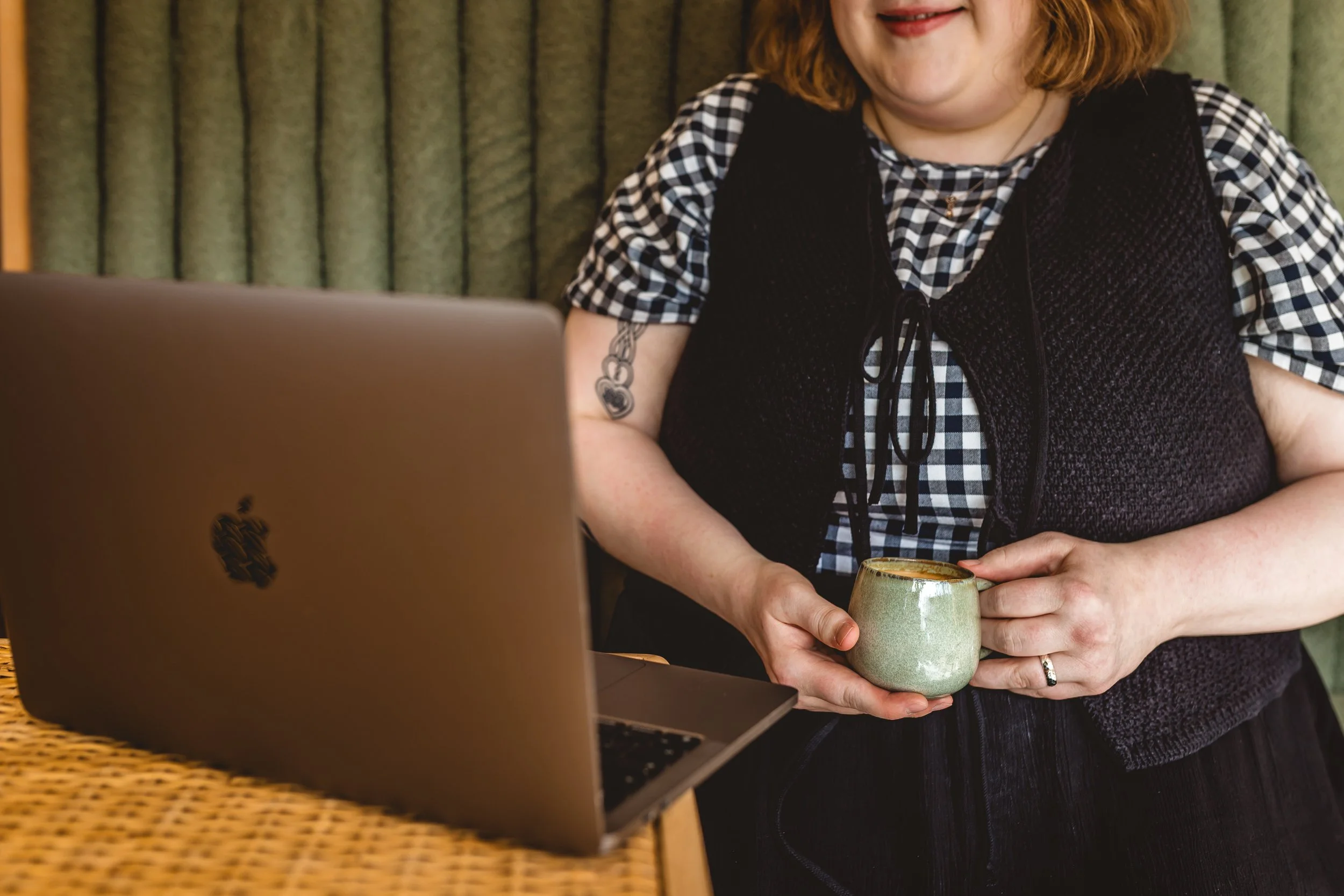 A woman with wavy brown hair and a tattoo on her arm sits at a table with a laptop, holding a ceramic mug.