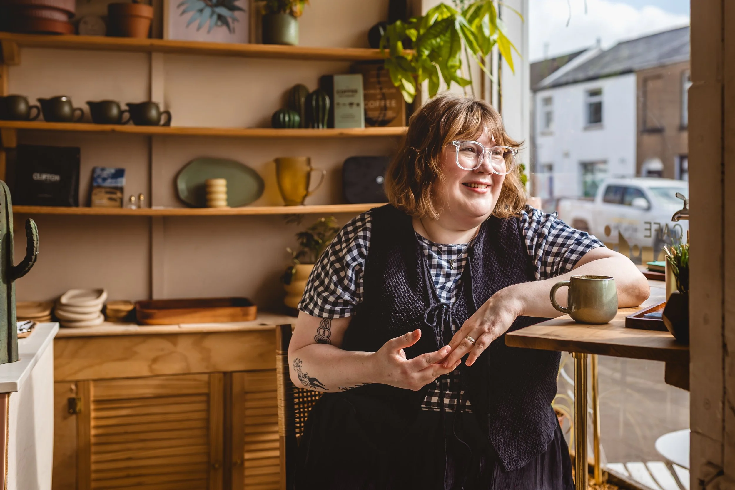 A woman with glasses and short brown hair sitting at a counter in a cozy cafe, smiling, with shelves of dishes and plants behind her, and a window showing neighboring buildings outside.