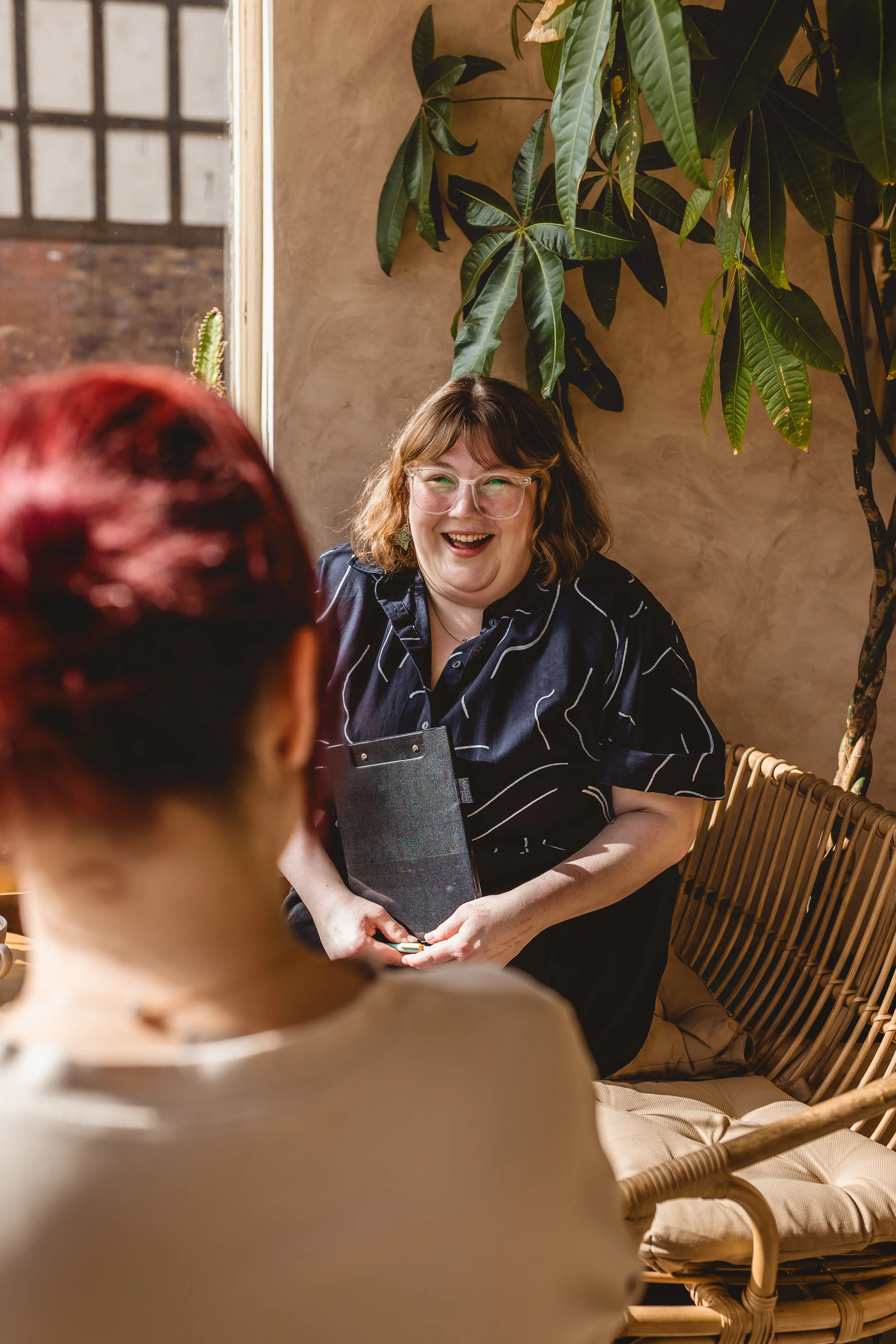 A woman with glasses, brown hair, wearing a black shirt with white stripes, smiling and holding a clipboard, sitting on a wicker chair in a room with large plants and a window.
