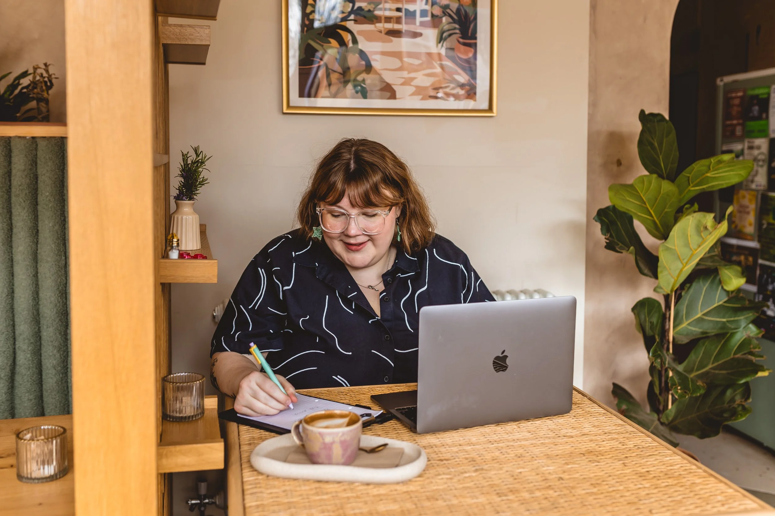 A woman with brown hair, glasses, and earrings working at a wooden desk with a silver Apple laptop, writing on a notepad with a green pen, surrounded by plants and decorative items in a cozy, well-lit room.