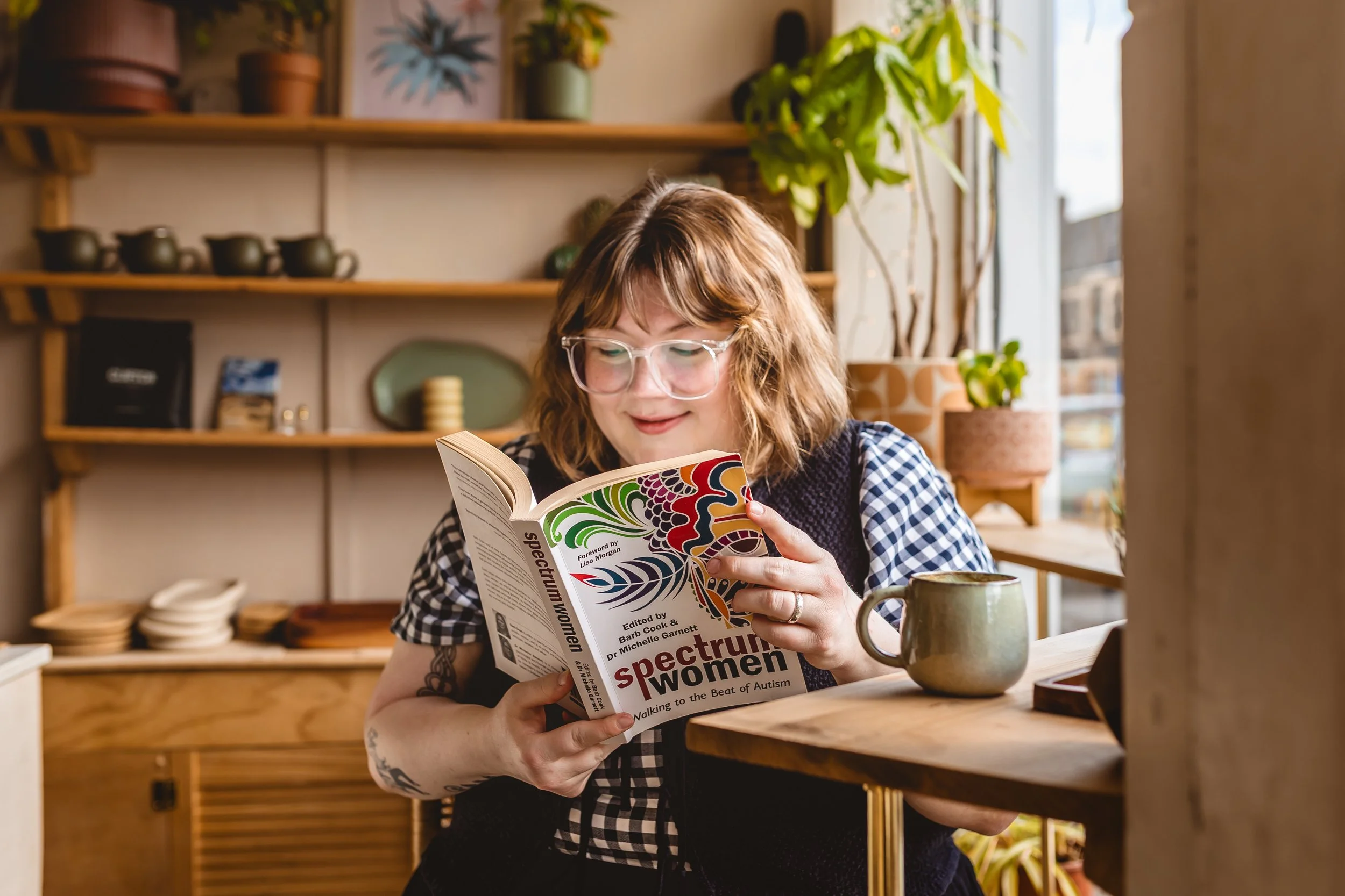 A woman with glasses and tattoos on her arm is sitting at a wooden table, reading a magazine about women with autism, in a cozy room with wooden shelves, potted plants, and dishes in the background.