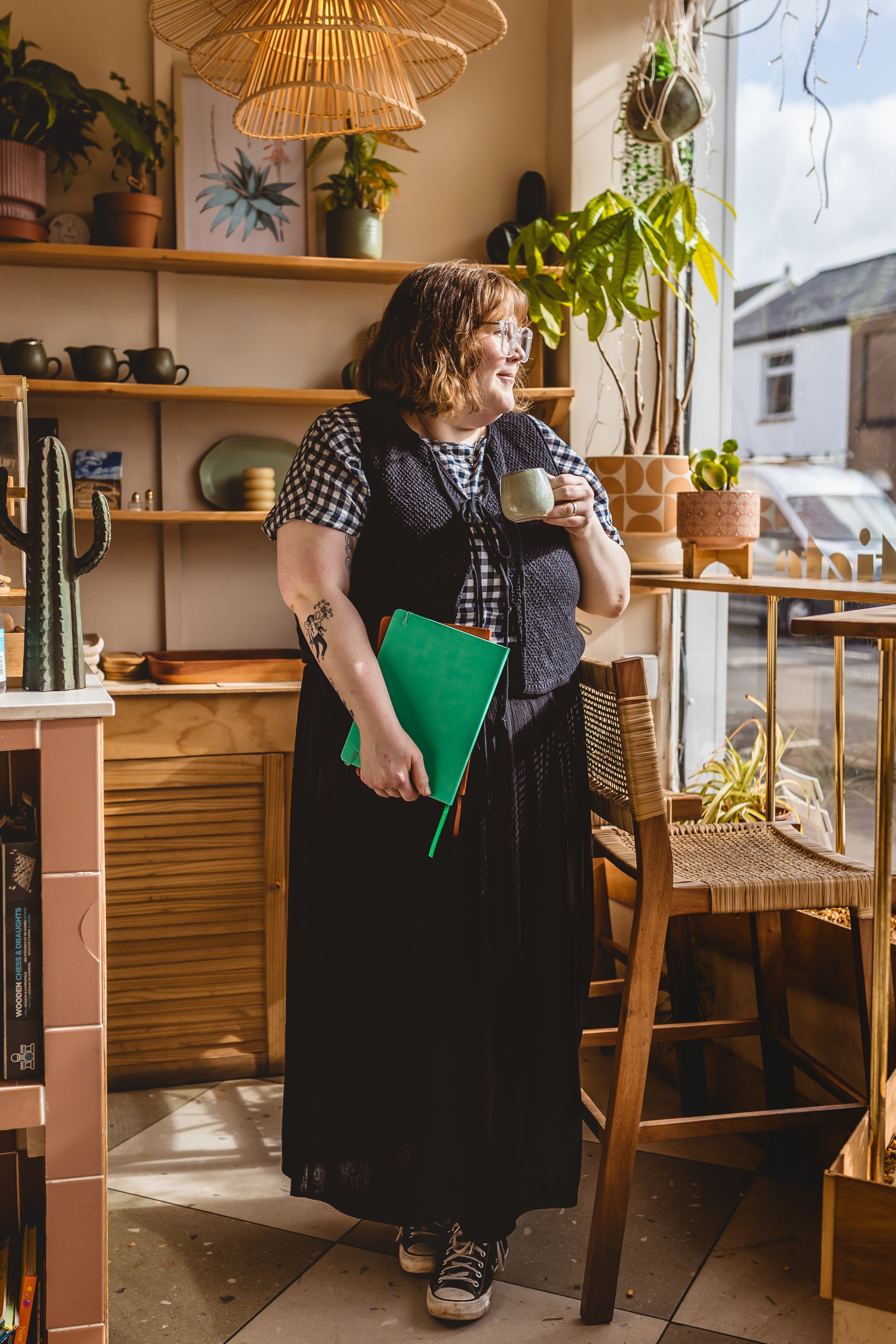 A woman with wavy brown hair and glasses holding a green folder, wearing a black and white checkered shirt and a black skirt, standing in a cozy indoor space with wooden shelves, plants, and decorative items, near a large window with sunlight.