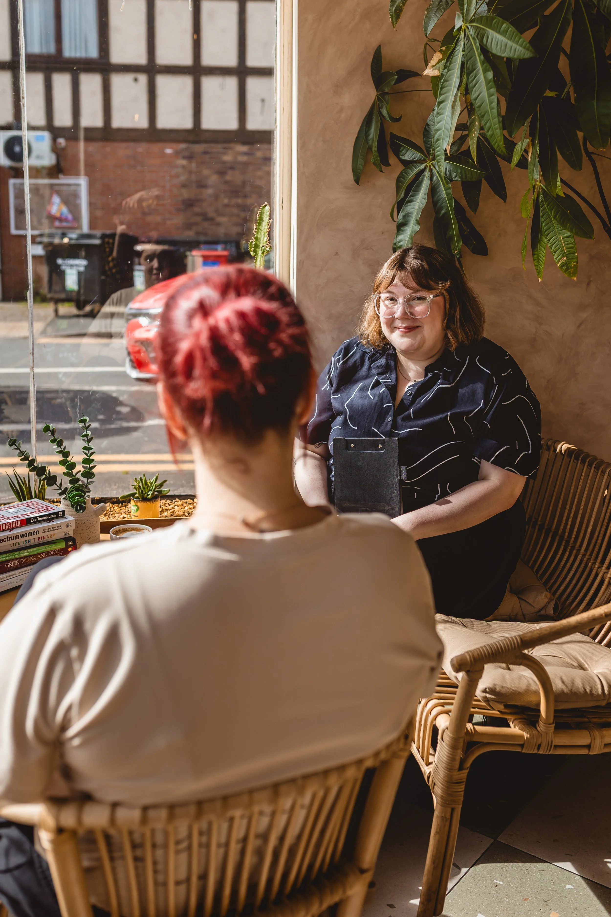 Two women are sitting indoors near a large window with a plant and books on the table. One woman is facing the other, and a woman with glasses and brown hair is smiling and looking the other woman.