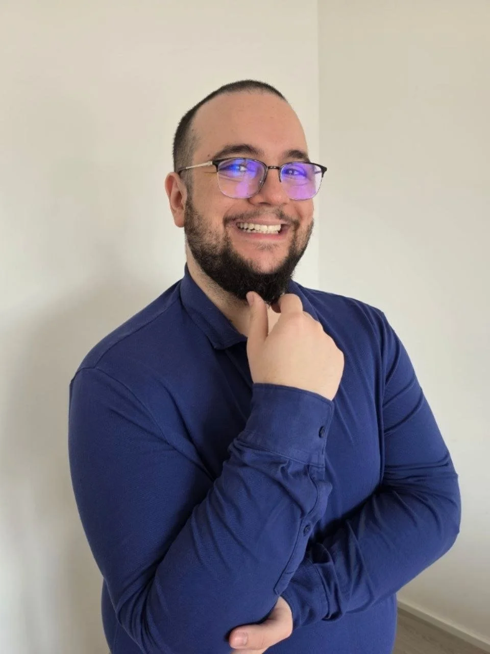 Ein lachender Mann mit Brille, dunklem Bart, kurz geschorenem Haar, in einem blauen Hemd, steht vor einer weißen Wand.