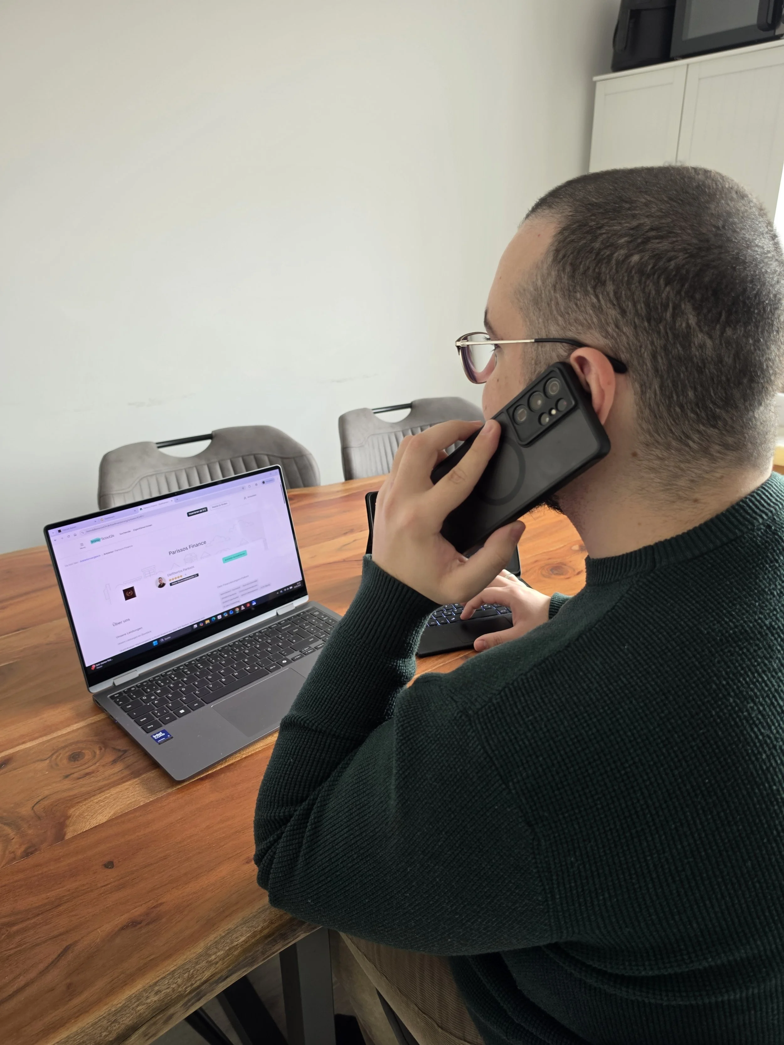 Ein Mann mit Brille sitzt an einem Holzkonferenztisch, spricht am Handy und benutzt einen Laptop.