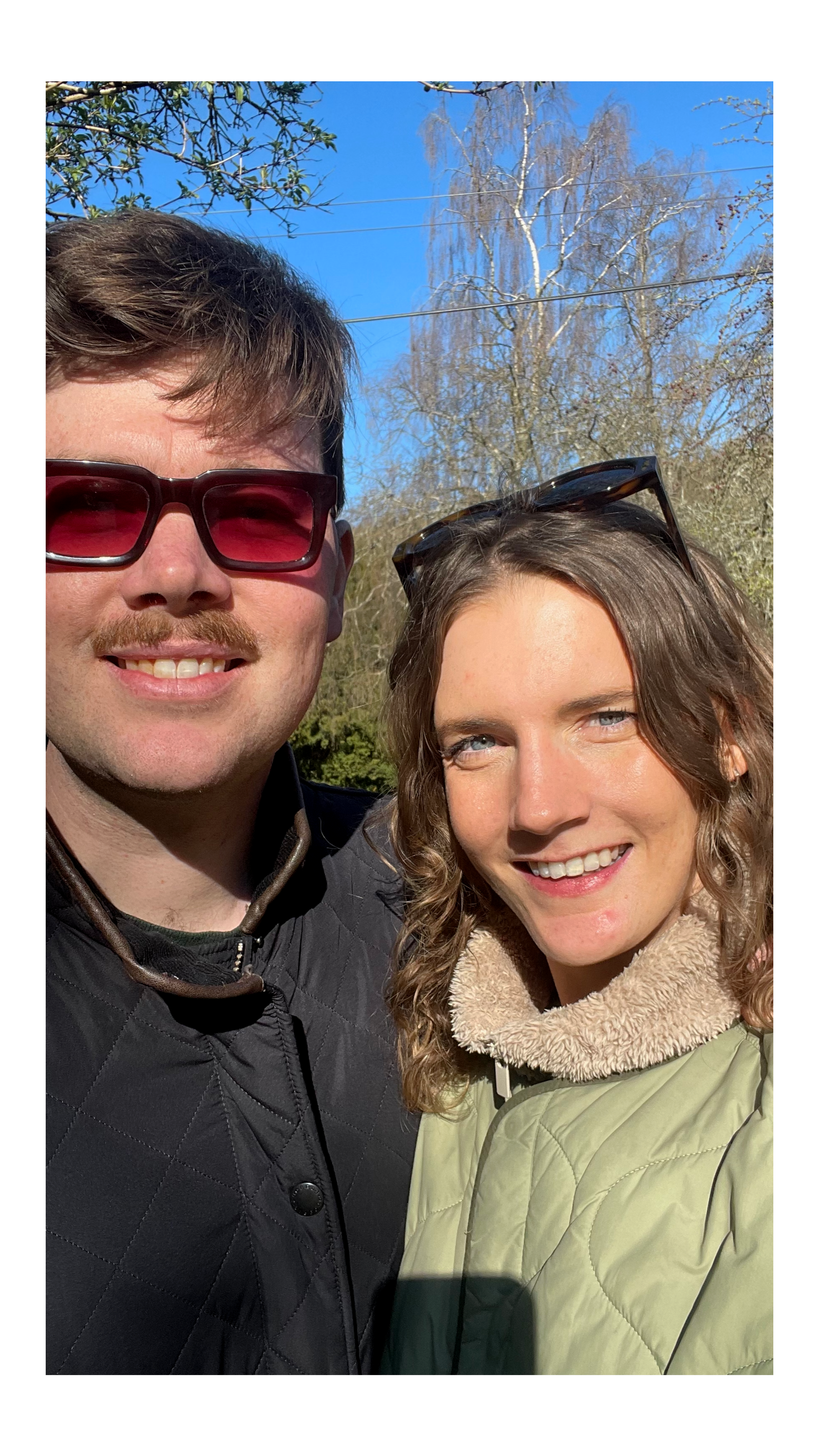 A man and woman smiling outdoors on a sunny day, with trees and a clear blue sky in the background.