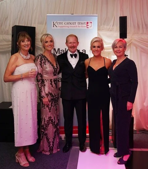 Five people standing together at a formal event, with a banner for Kent Cancer Trust in the background. Four women and one man, dressed in elegant attire, smiling for the photo.