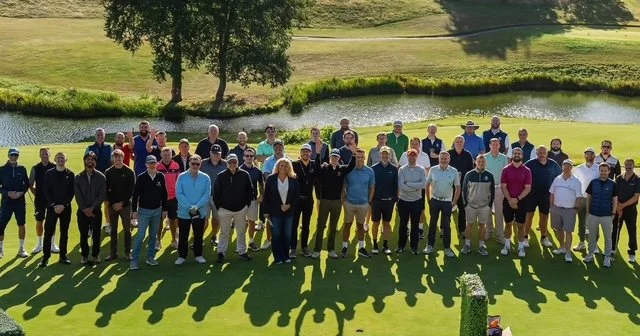 Group of people standing on a golf course near a small pond with trees and grass in the background.
