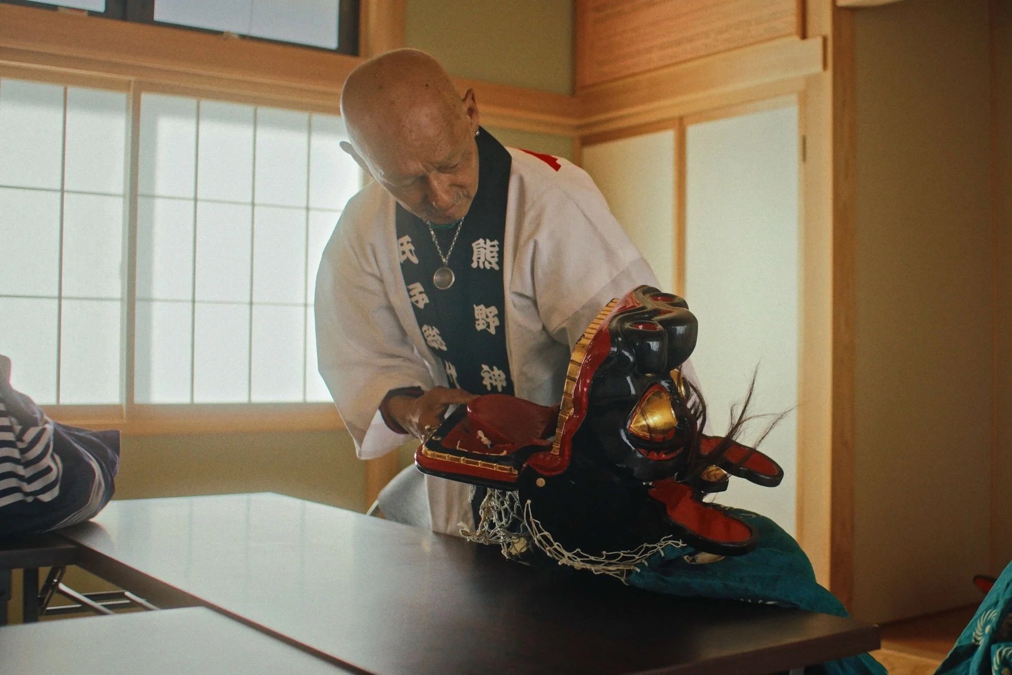 A bald man dressed in a traditional Japanese attire, holding a samurai mask and armor, either preparing for or engaged in a cultural activity or ritual inside a room with wooden paneling and shoji screen windows.