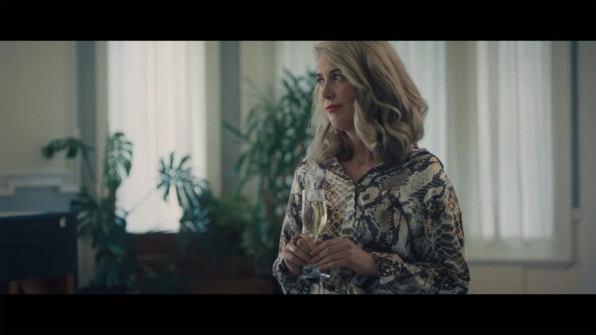 A woman with blonde wavy hair wearing a snakeskin patterned blouse holds a glass of champagne in a well-lit room with green plants and light-colored curtains in the background.