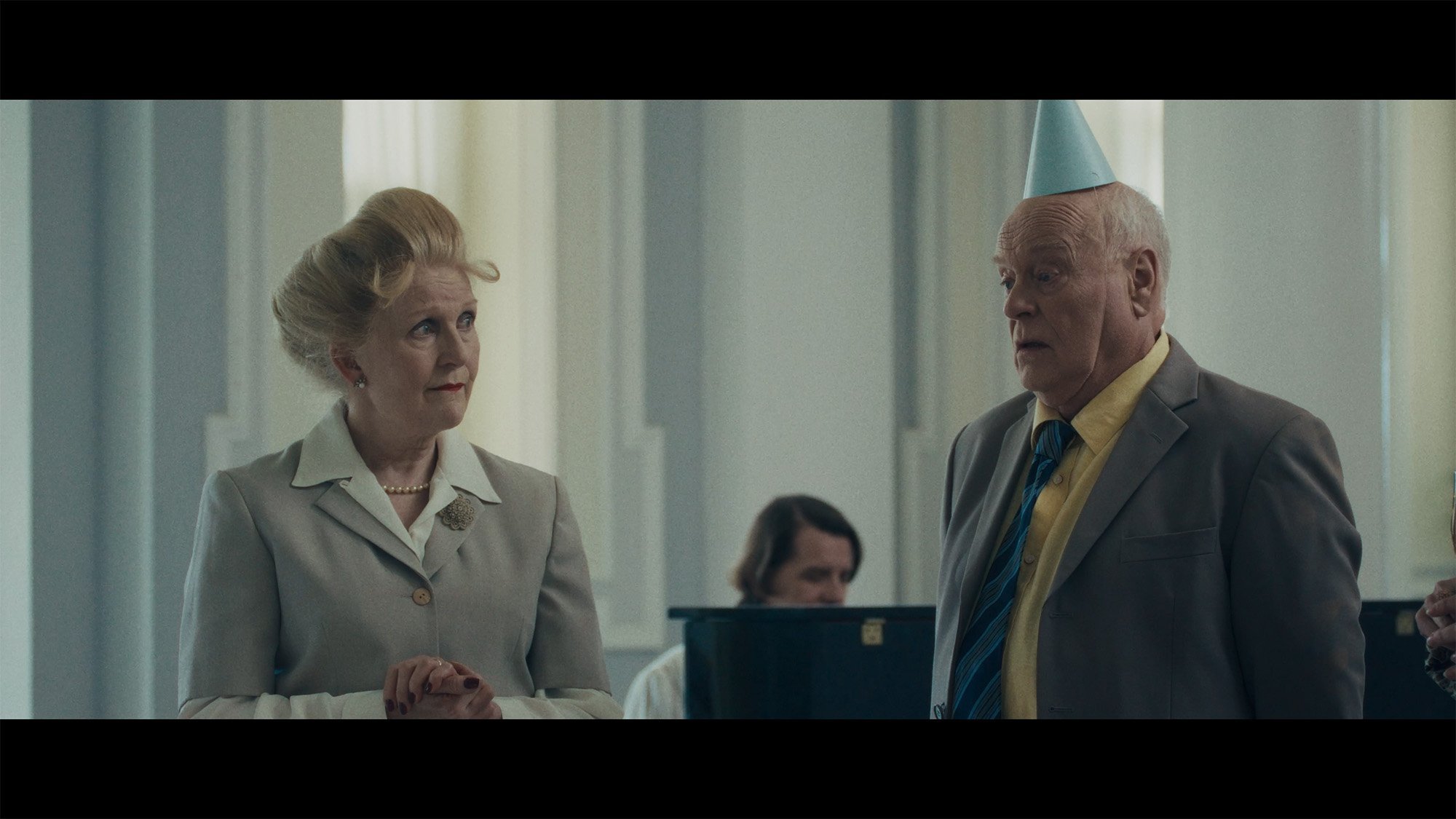An elderly woman and man standing indoors at a formal event, dressed in vintage clothing. The woman has styled blonde hair, wears a light-colored suit with a pearl necklace, and the man has white hair, wears a gray suit, a yellow shirt, and a party h