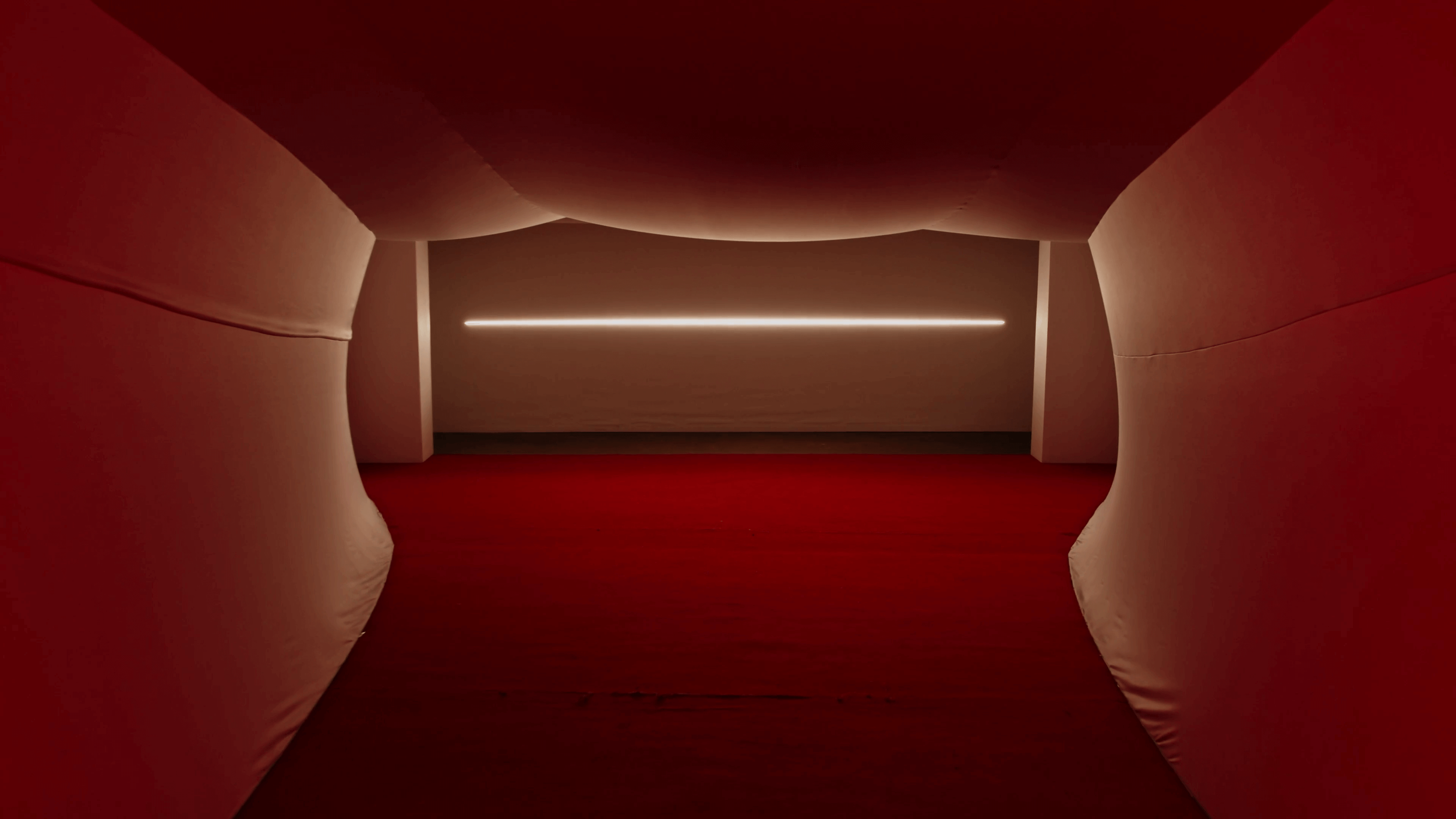 Interior view of an art installation by Es Devlin at 180 Strand. A corridor with curved red walls, red flooring, and a minimalist horizontal light fixture on the wall.