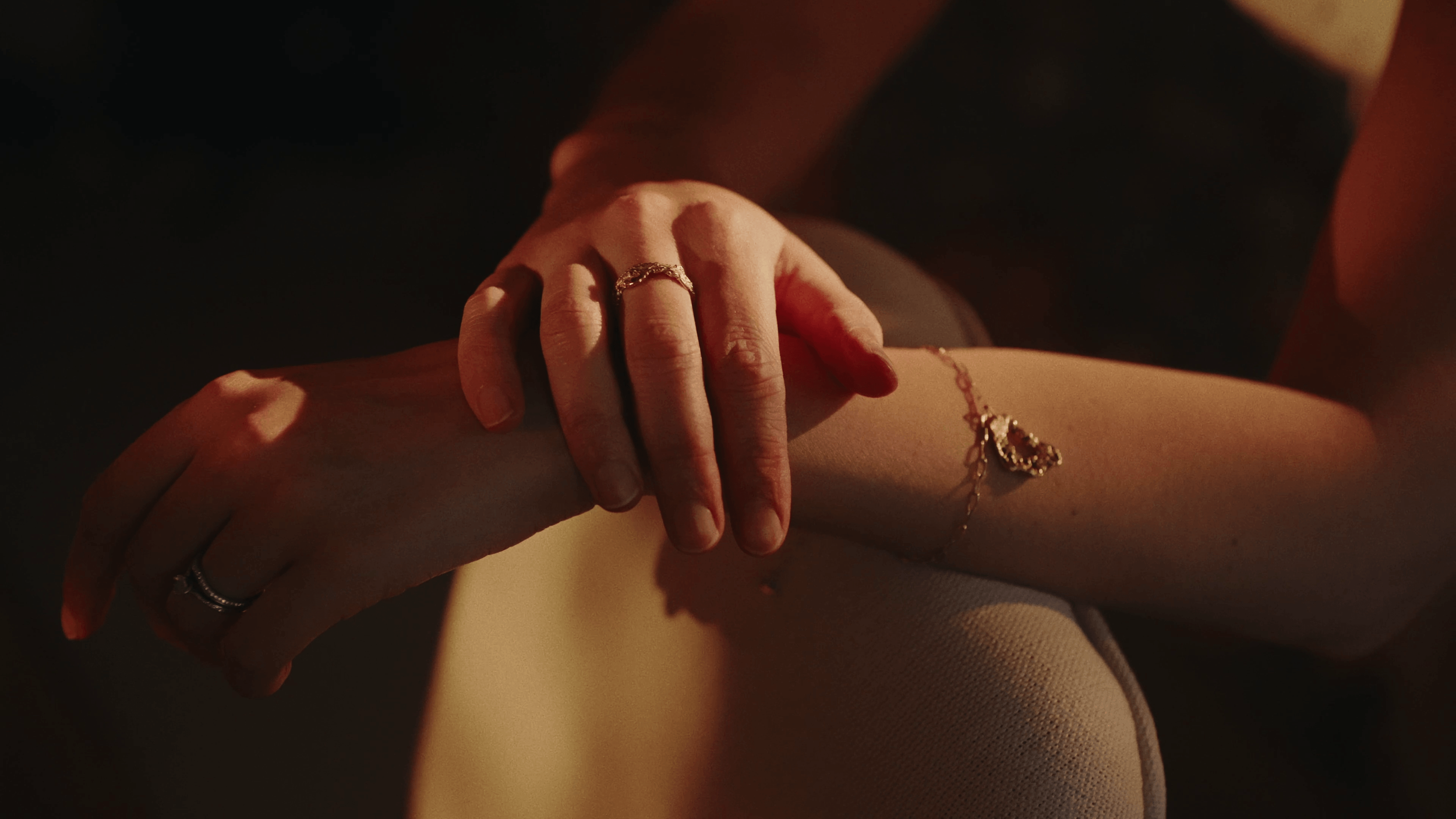 Close-up of two hands, one resting on the other's arm, with jewelry including rings and a bracelet, in a dimly lit setting.