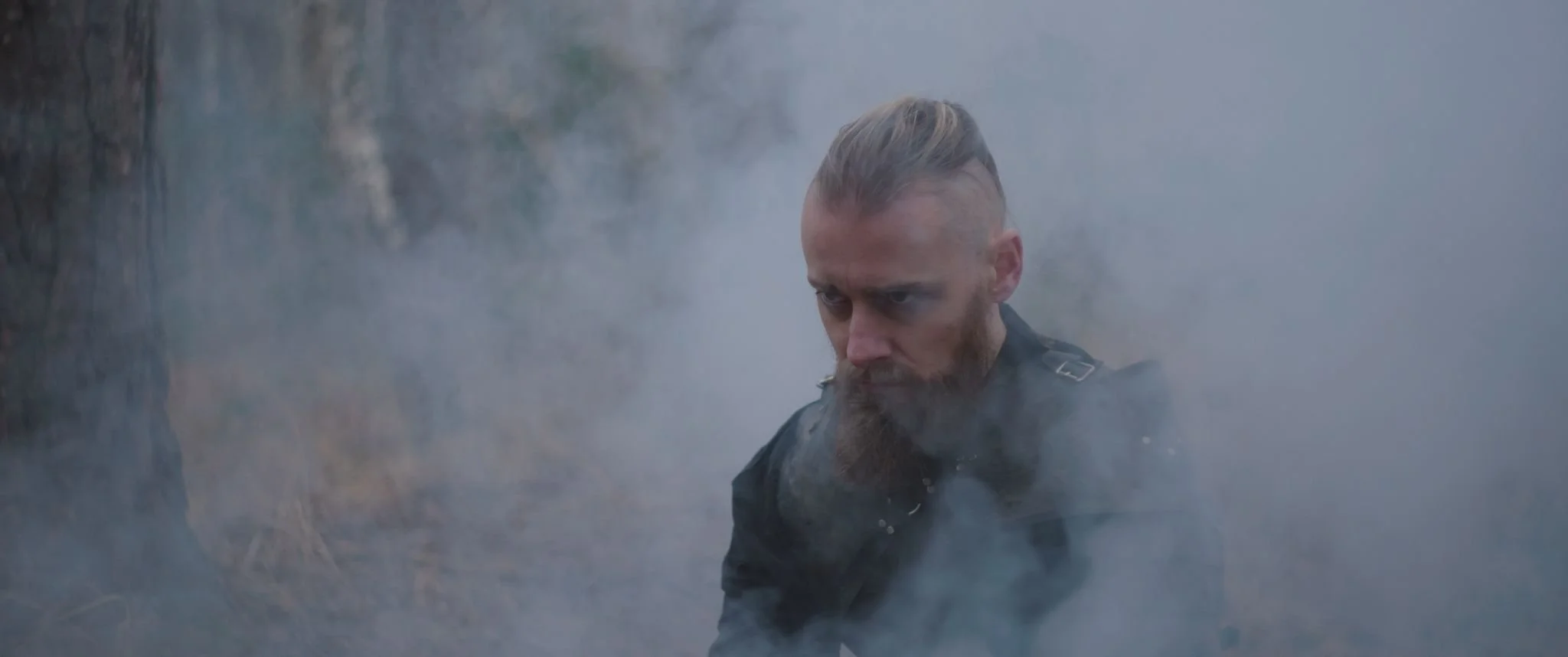A viking man with a partial beard and stylized hair, dressed in viking armour, looking intense amidst a smoky environment outdoors.