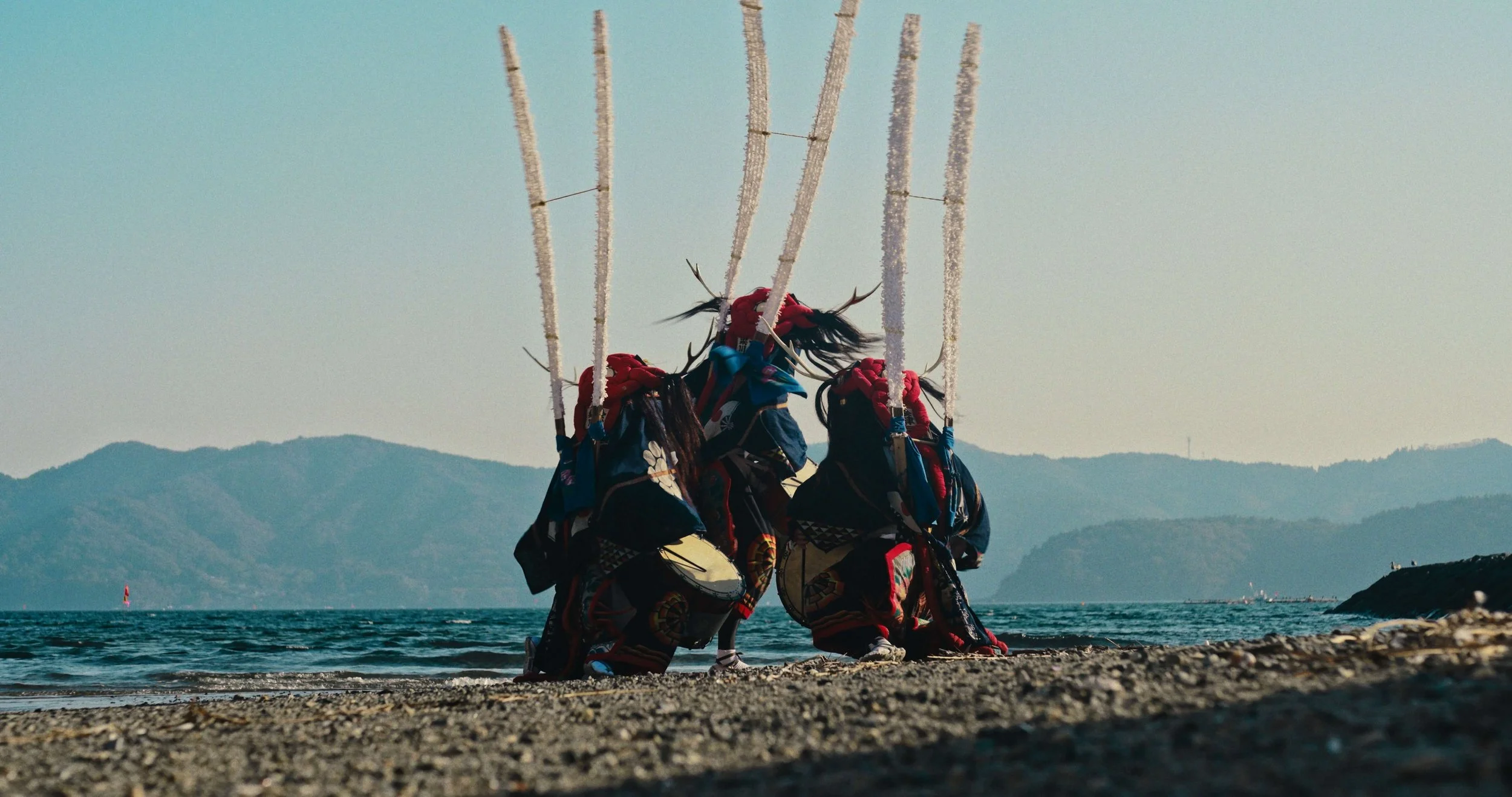 A still image from Stronger than the Wave, a documentary film by Robbie McKane, featuring three dancers in Shishi-Odori garb, a popular folk dance from the Tohoku region performing on a beach with mountains in the background and a boat in the water.