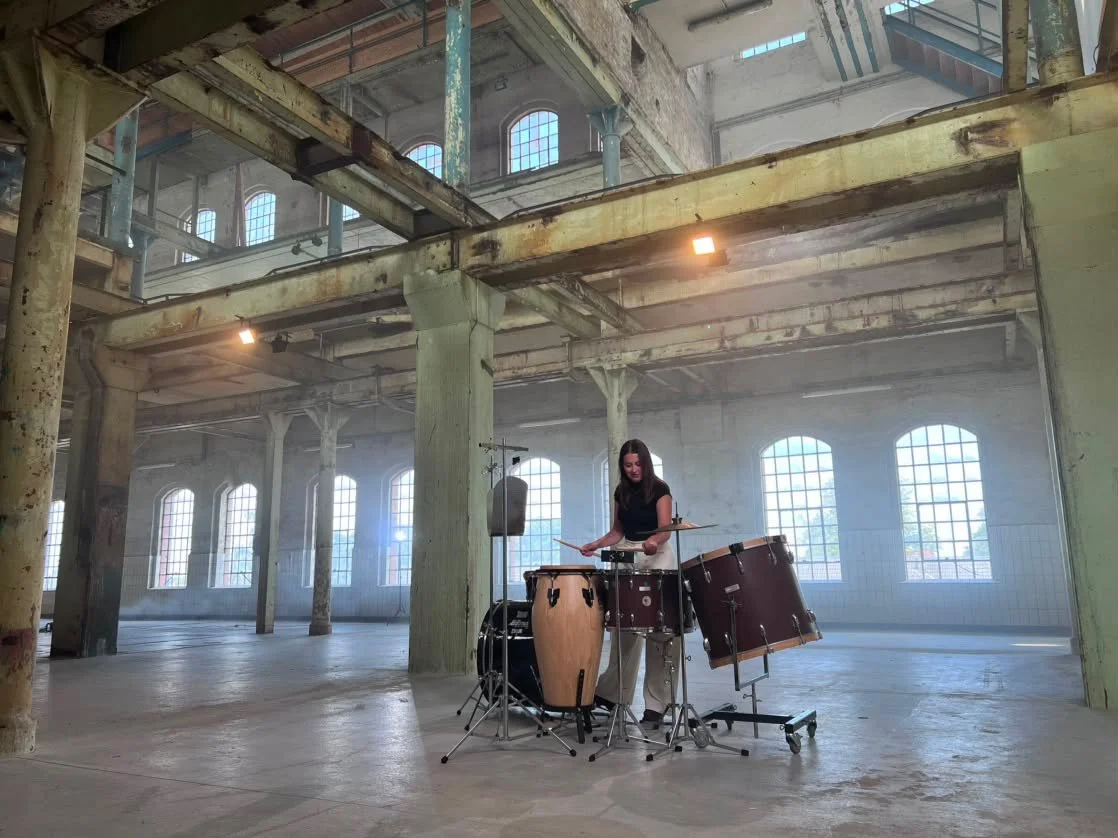 A woman playing drums in an empty, industrial-style space with large windows and exposed beams.