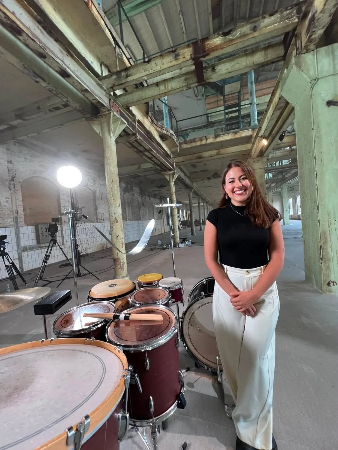 A woman in a black top and white pants standing next to a drum set in an industrial, exposed brick space with photography equipment.