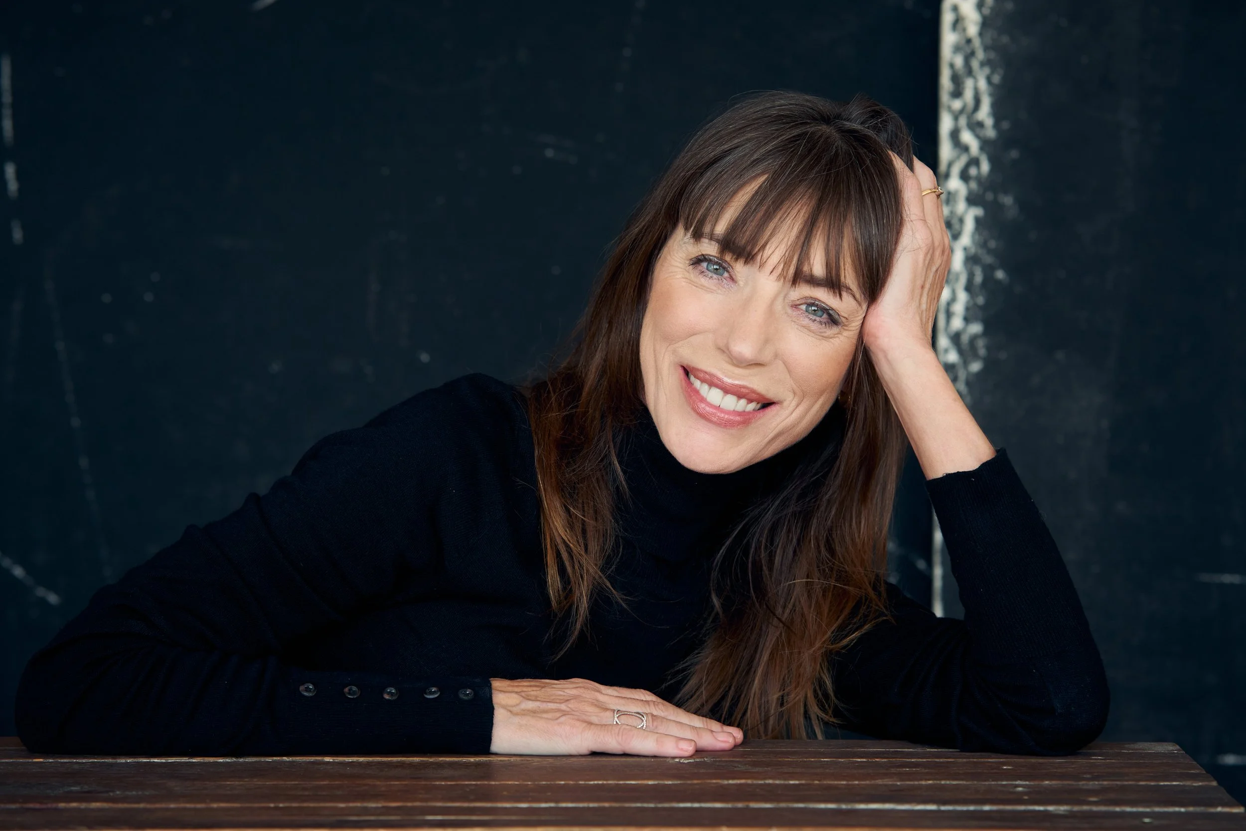Mujer con cabello castaño y ojos claros, sonriendo, apoyada sobre una mesa de madera, con un fondo oscuro.