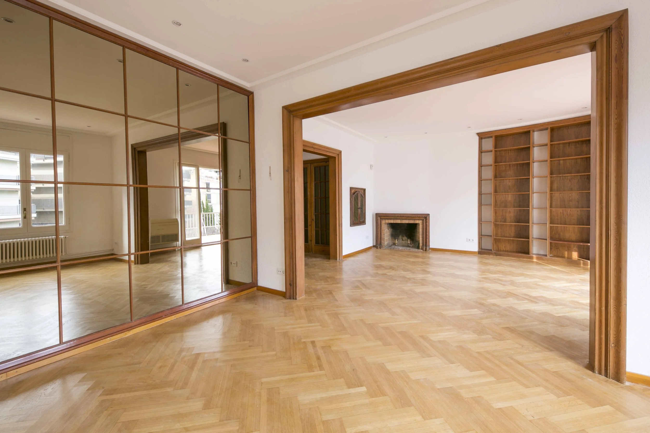 Sala de estar con paredes blancas, pisos de madera, chimenea y estantería de madera. Una pared con ventanas y un espejo grande de madera separado por marcos de madera.