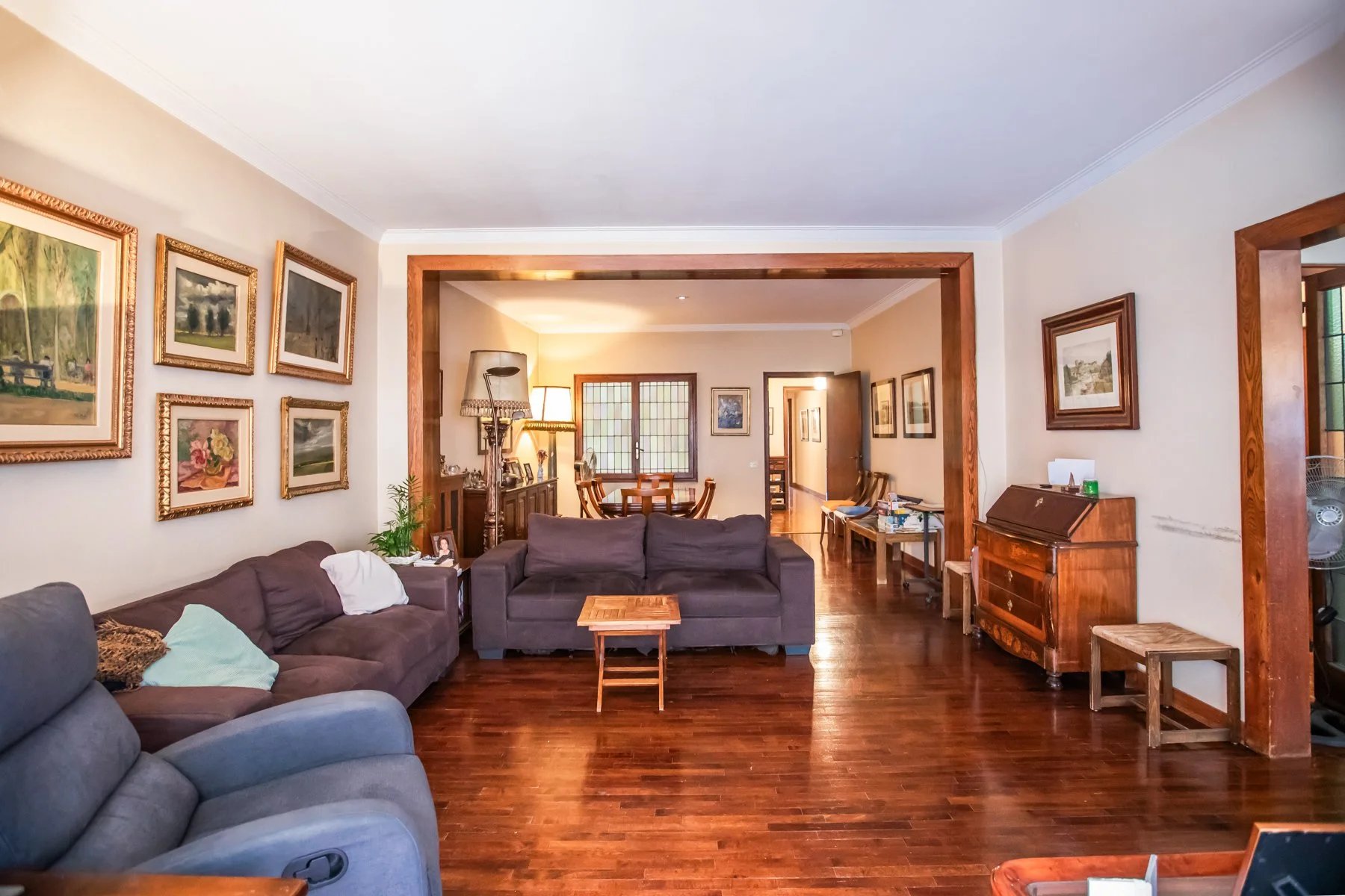 Sala de estar con sillones grises, cuadros en las paredes, piso de madera, y una vista hacia el comedor y pasillo en el fondo.