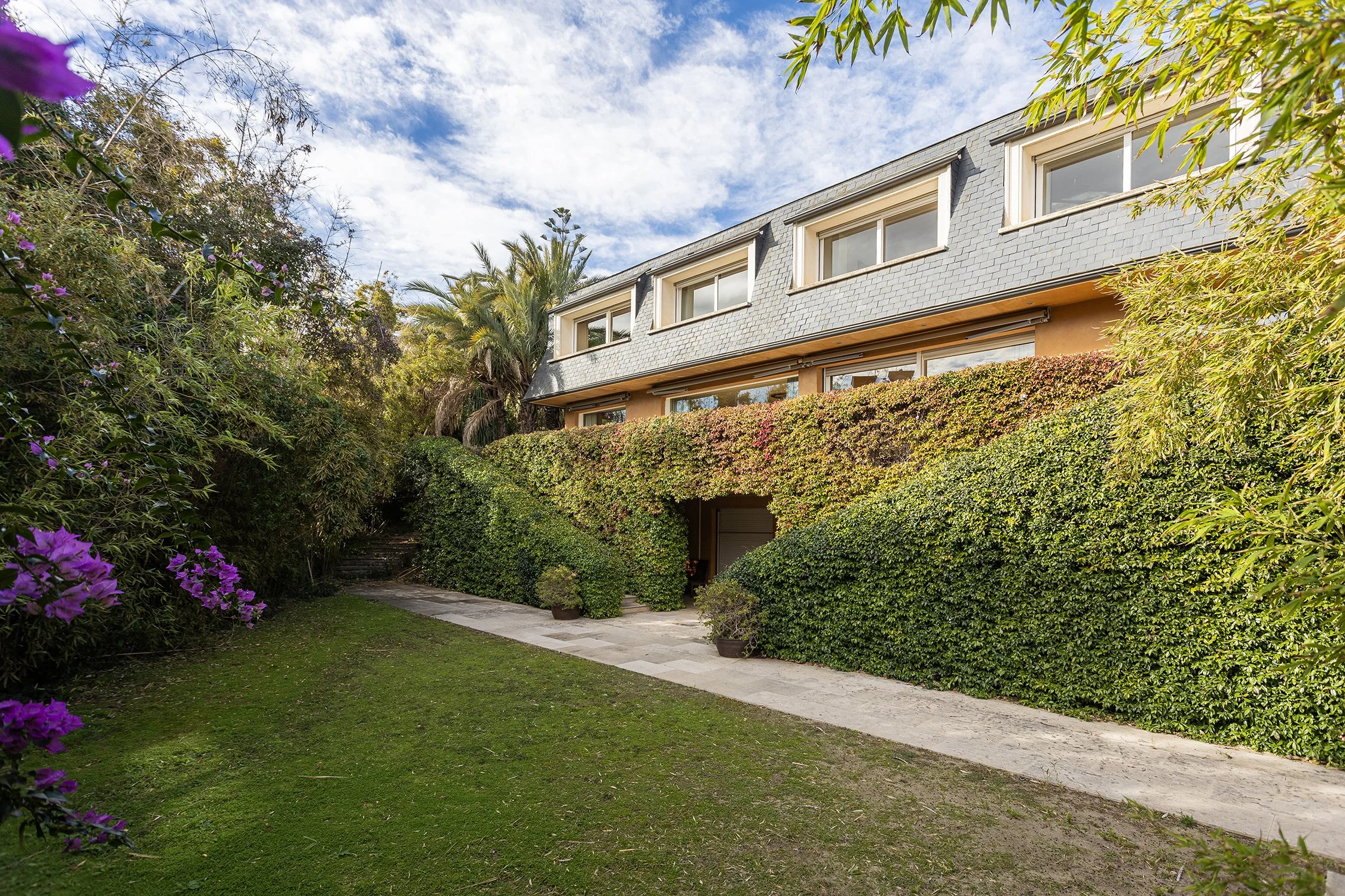 Casa moderna con ventanas grandes, rodeada de vegetación y árboles, en un día soleado con cielo azul y nubes.