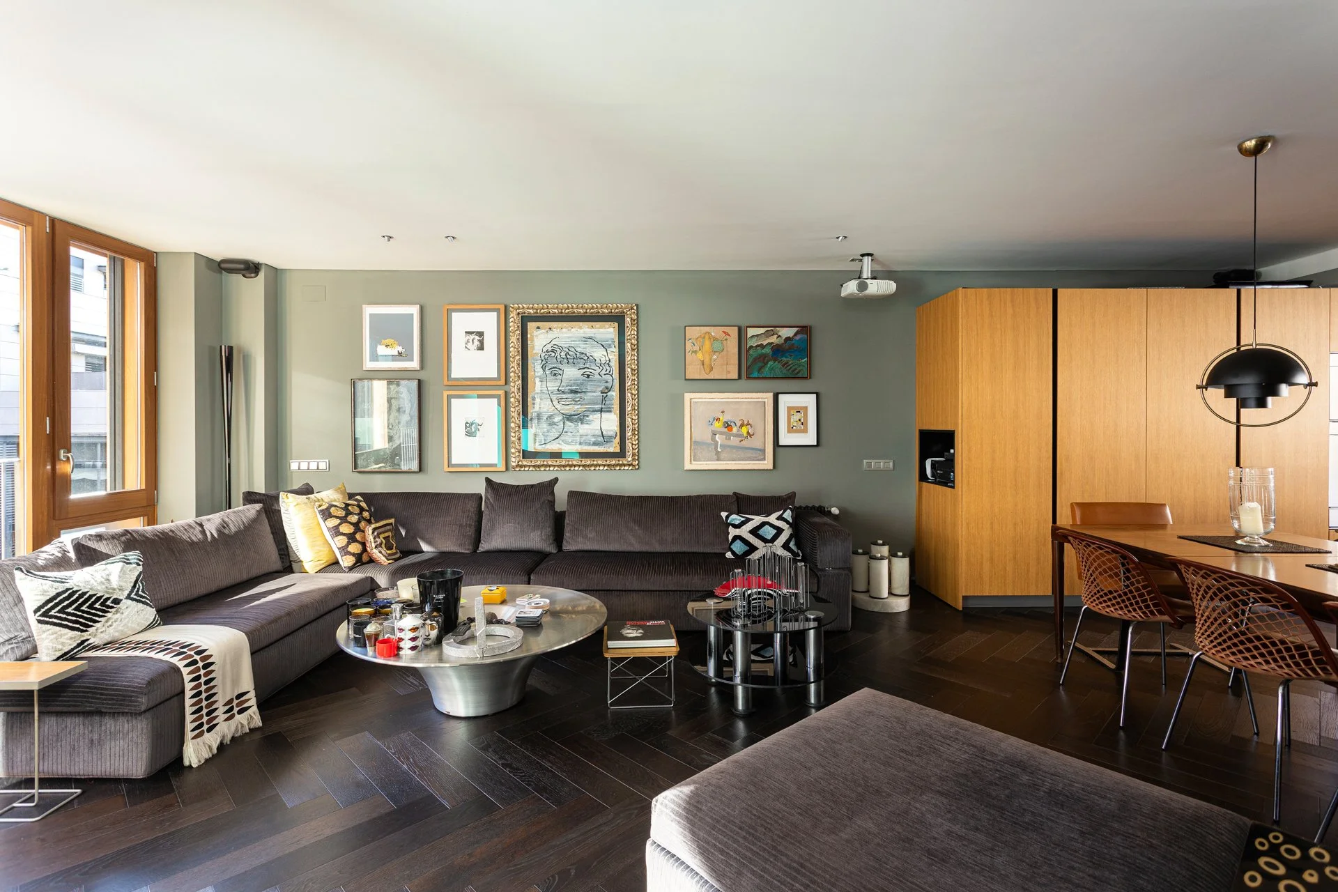 Sala de estar moderna con sofá oscuro, mesa de centro con objetos decorativos, paredes con varias pinturas y cuadros, luminarias colgantes, ventanas amplias con marcos de madera, y área de comedor con sillas de diseño.