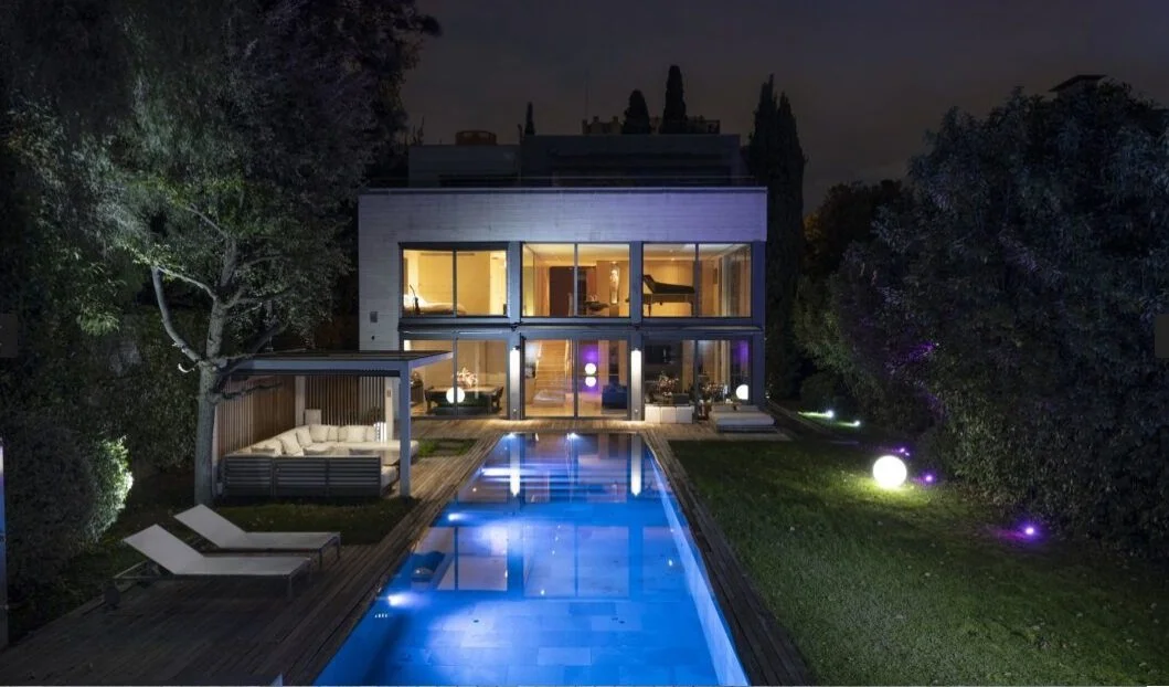 Imagen de una casa moderna de varias plantas, vista nocturna, con piscina iluminada y mobiliario exterior, rodeada de árboles y áreas verdes.
