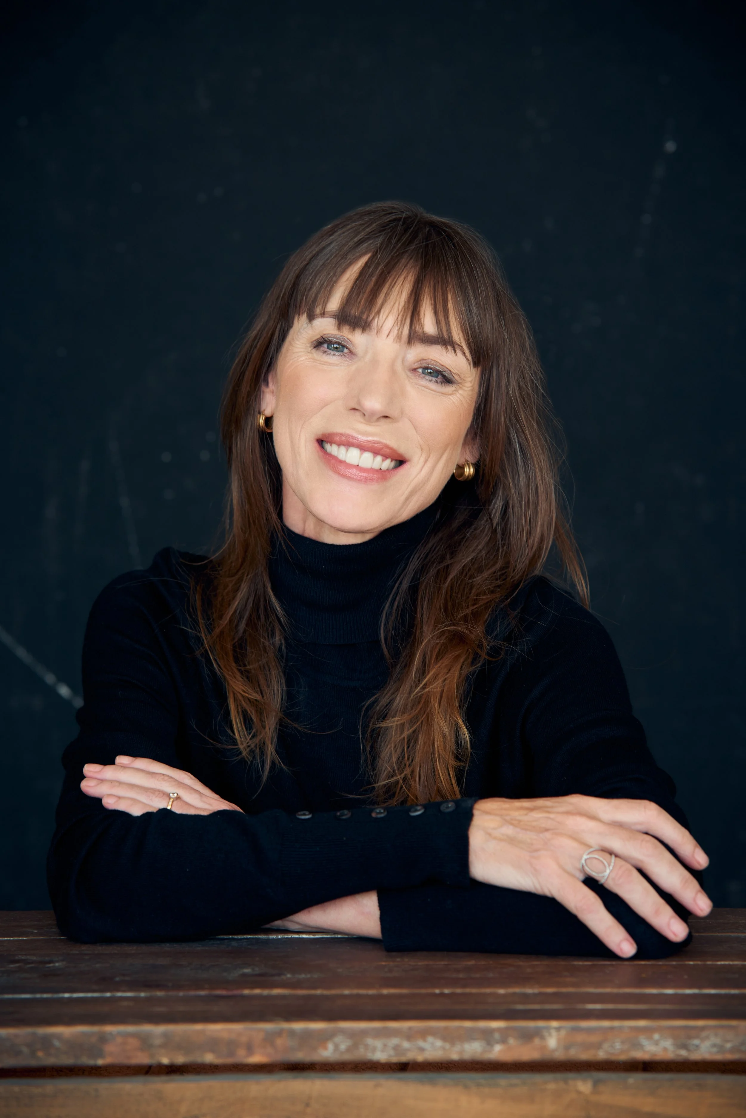 Mujer de cabello castaño claro y ojos azules, sonriendo, usando un suéter negro, con fondo oscuro y una mesa de madera en primer plano.