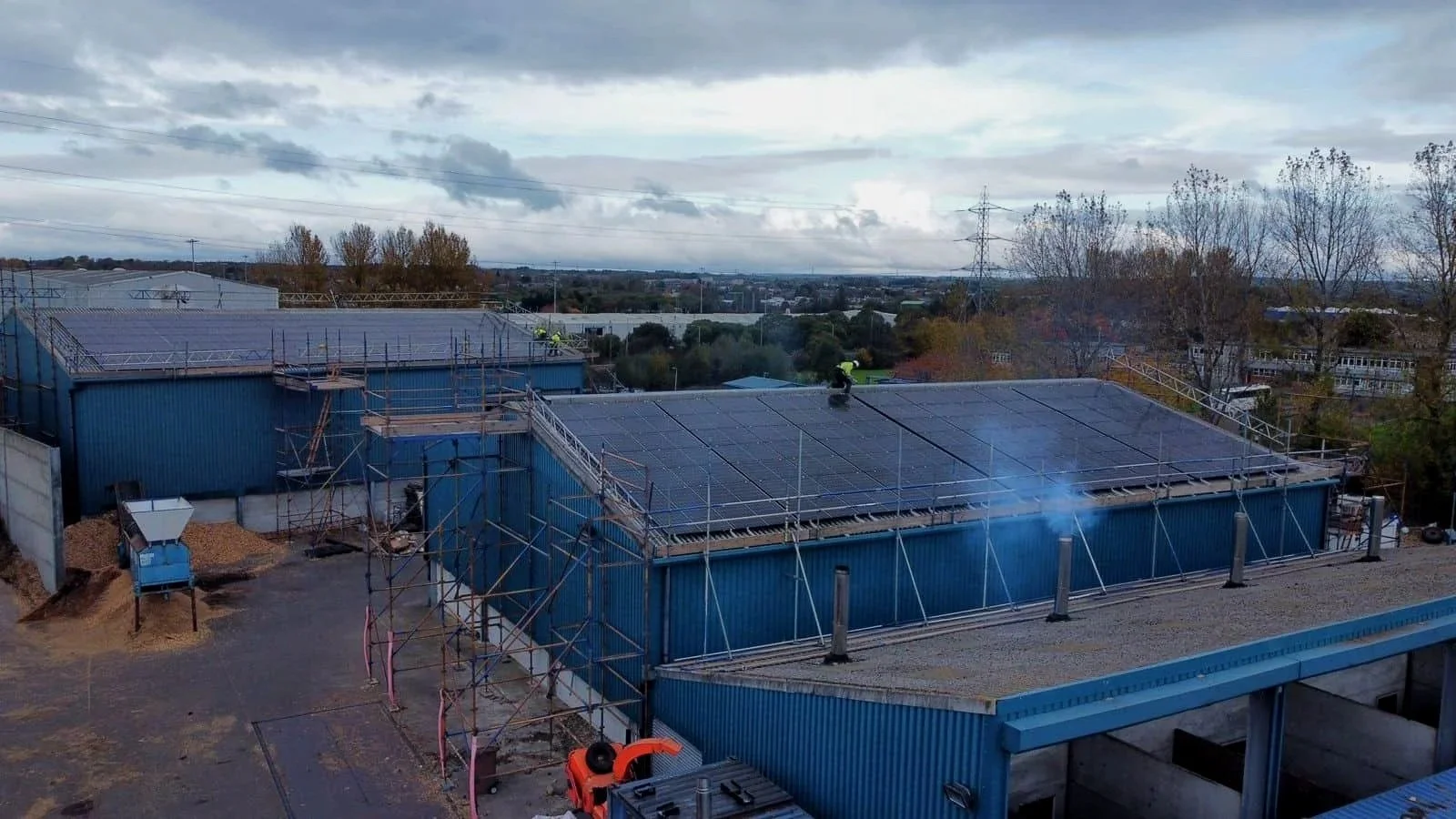 Construction workers on blue industrial building roof installing solar panels, surrounded by scaffolding, with trees and cloudy sky in background.