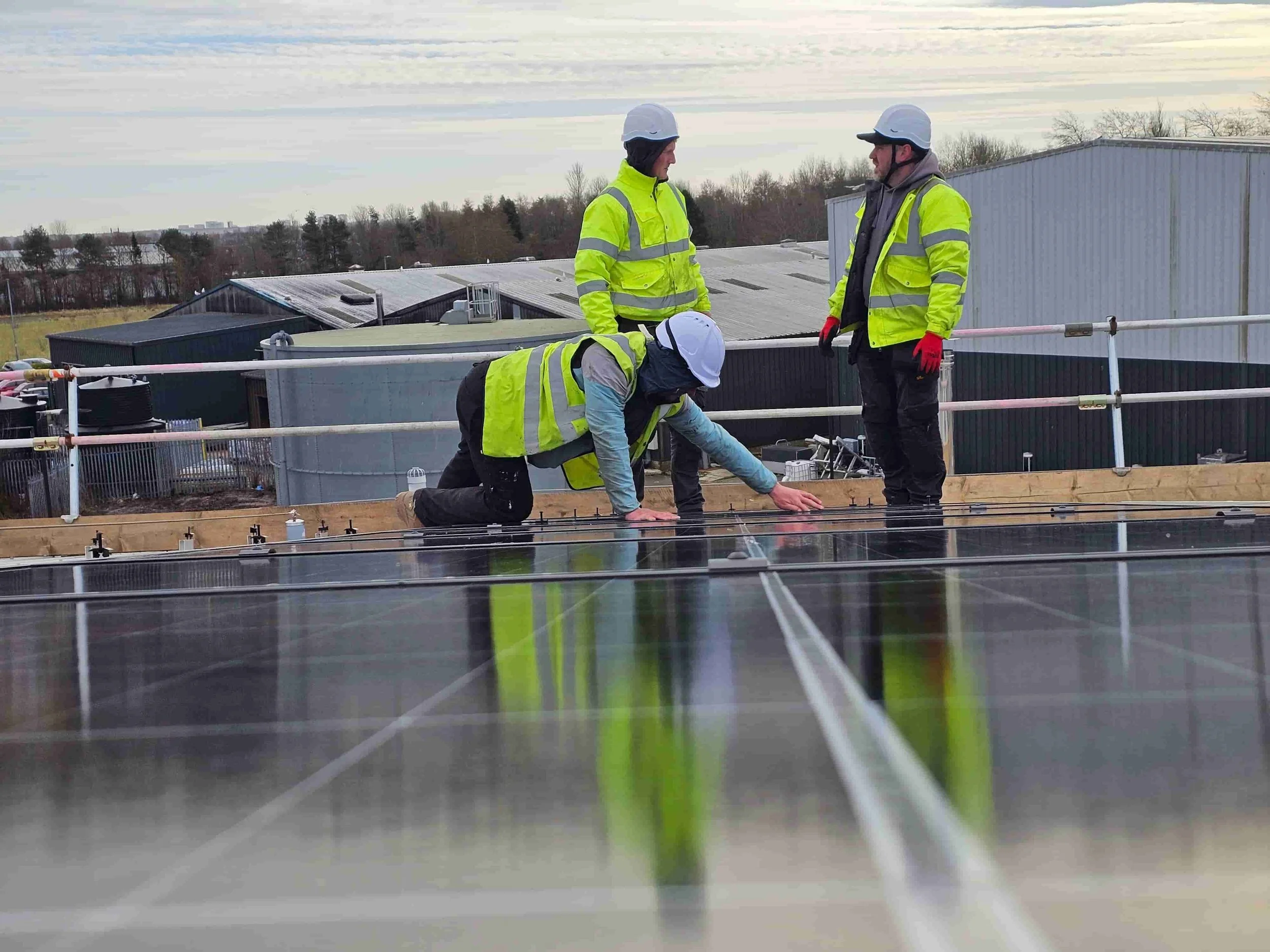 Three workers in safety vests and helmets inspecting solar panels on a rooftop during daytime.
