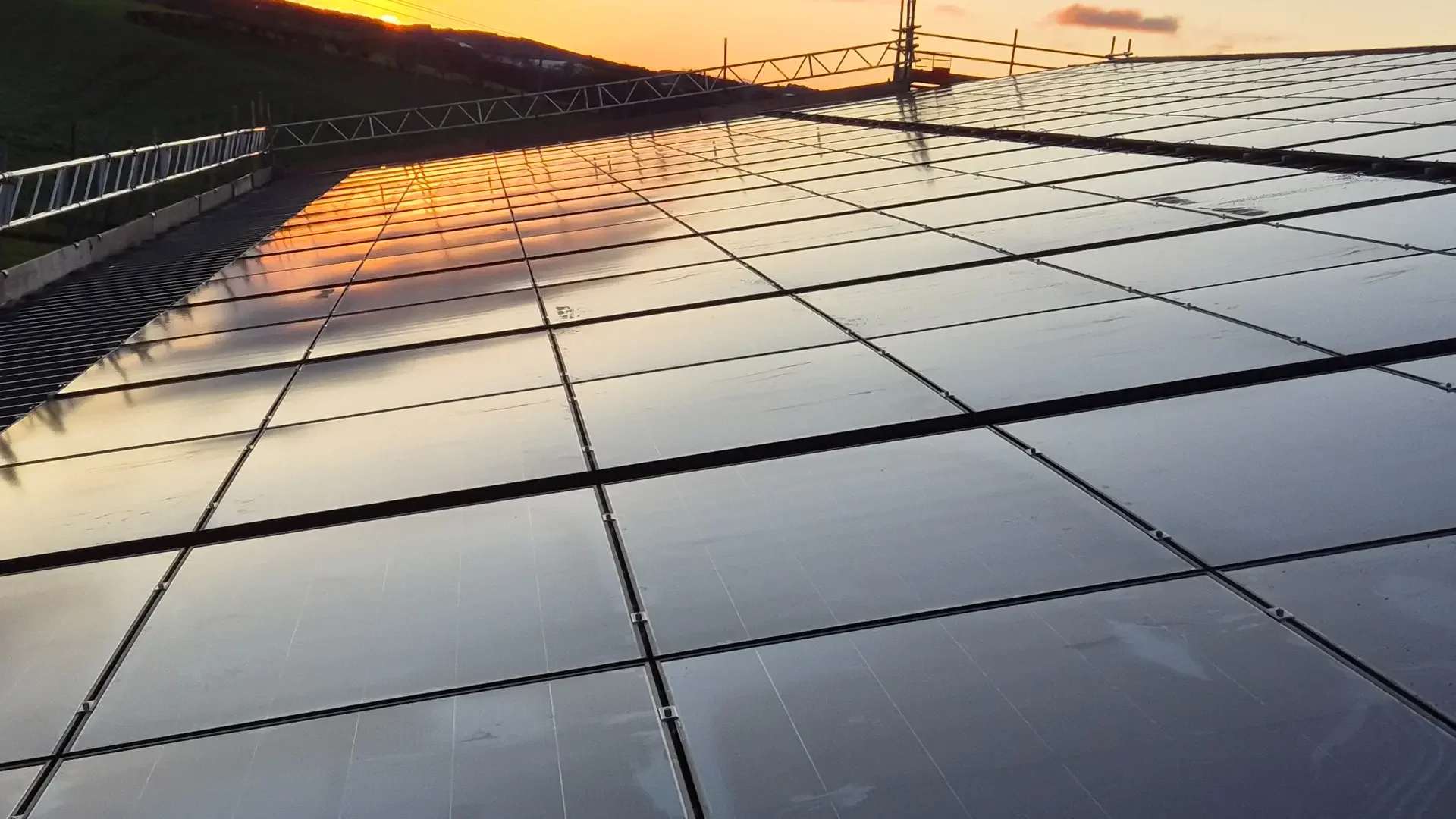 Sunset reflecting on multiple solar panels mounted on a rooftop with a railing alongside, with hills and a partly cloudy sky in the background.