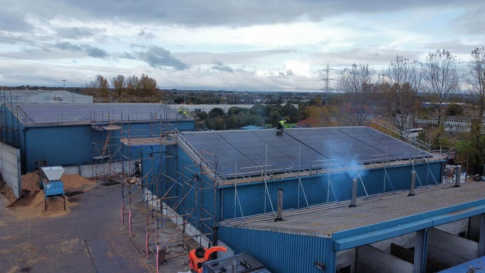 Workers installing solar panels on the roof of a blue industrial building with scaffolding around it, in a semi-rural area under a cloudy sky.