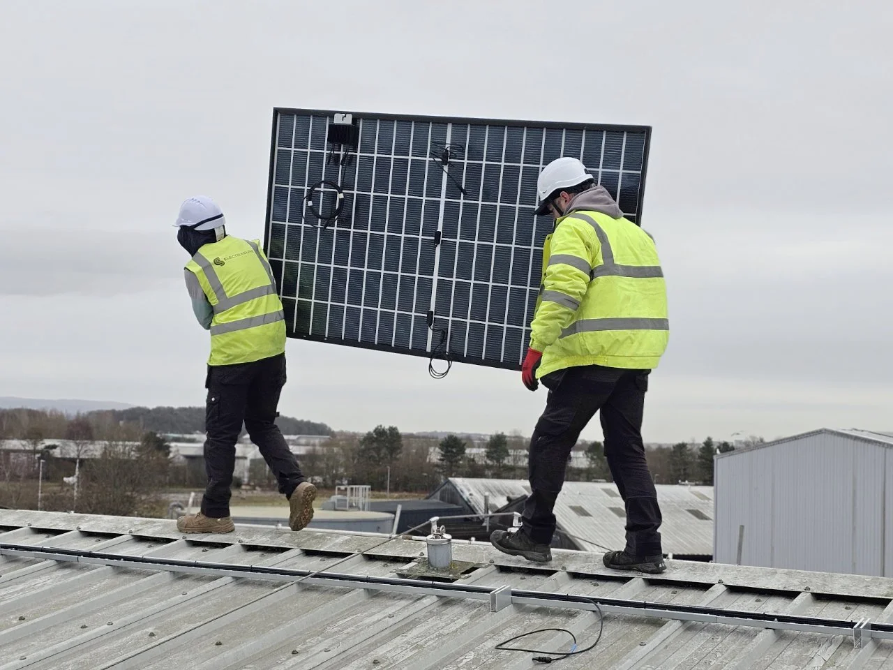 Two workers wearing yellow safety vests and white helmets installing or maintaining a large solar panel on a rooftop.