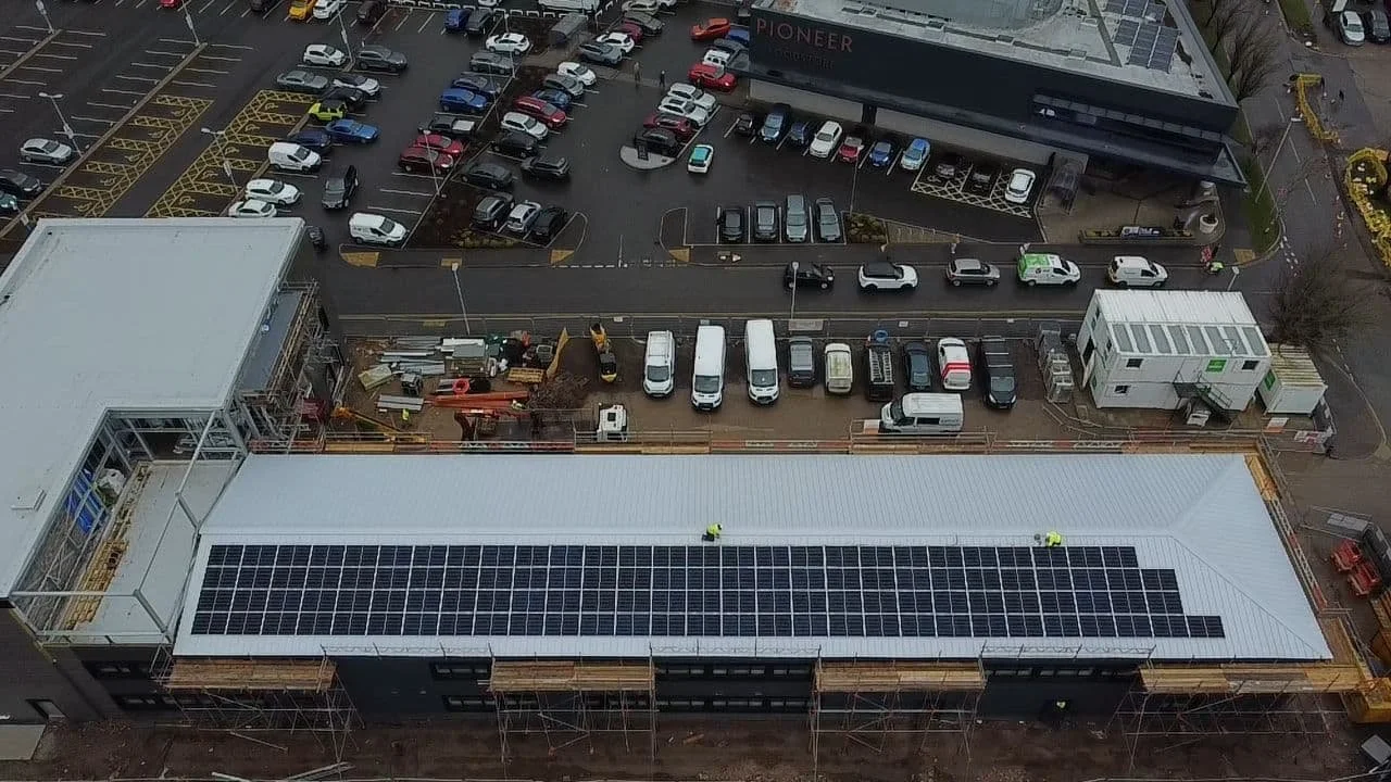Aerial view of a building under construction with solar panels installed on the roof, surrounded by parked cars and construction materials.