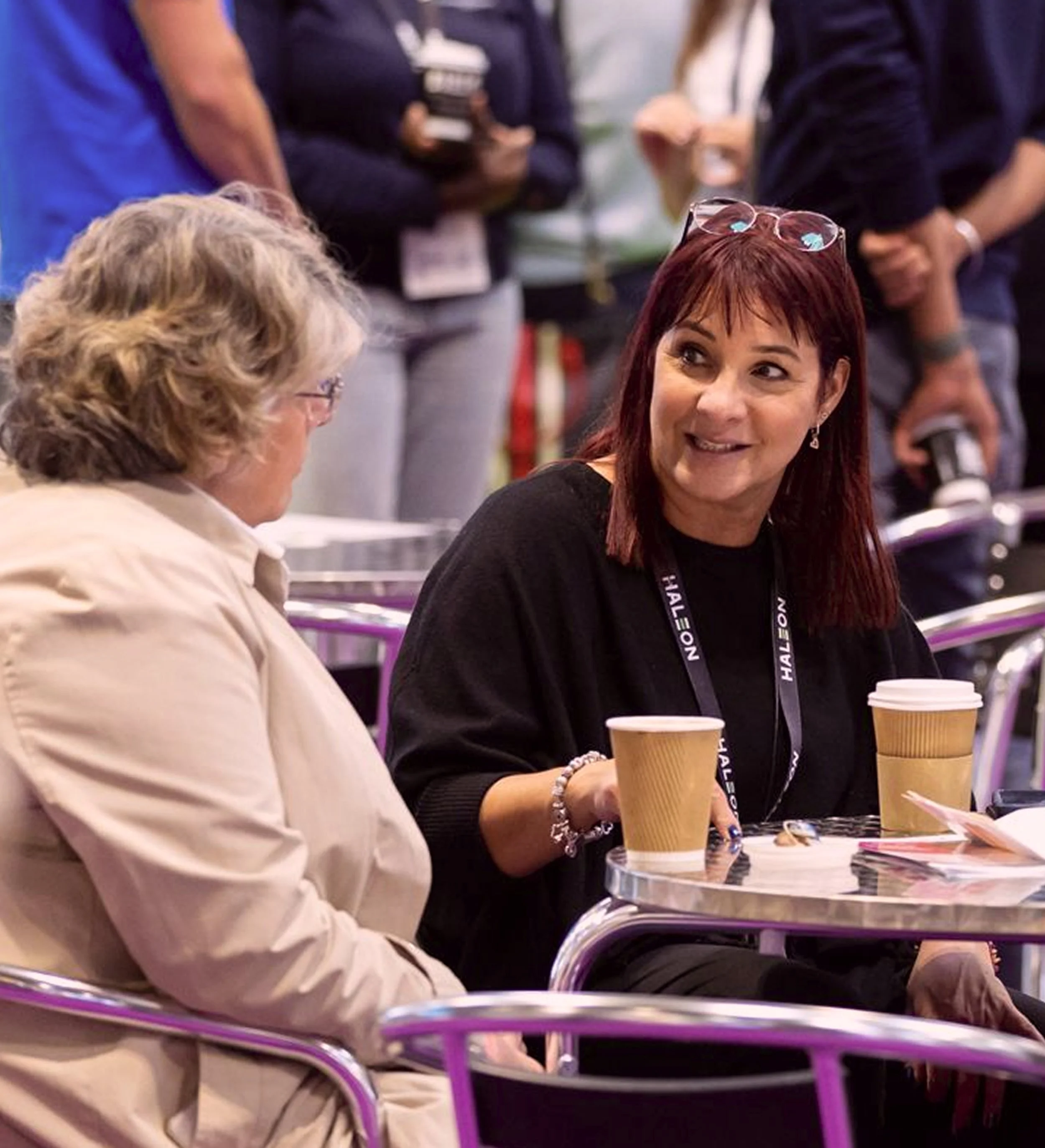 Two women sitting at a table with coffee cups, engaged in a conversation at a conference or event, with people standing in the background.