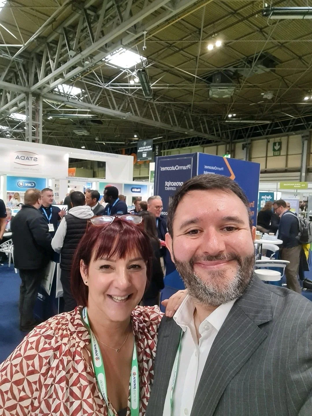 Selfie of a man and woman at a trade show or conference, with several people in the background engaging in discussions, booths, and display signs.