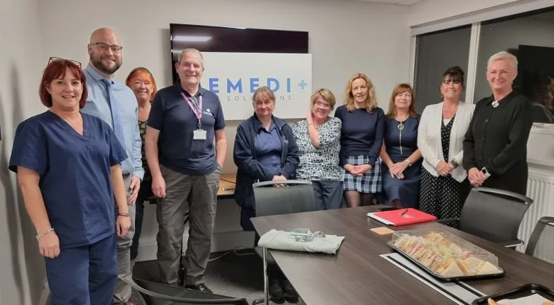 Group of nine people, including medical staff, standing in a conference room in front of a screen displaying MEDI+ SOLUTIONS" logo, with a table in front holding a tray of sandwiches and some documents.