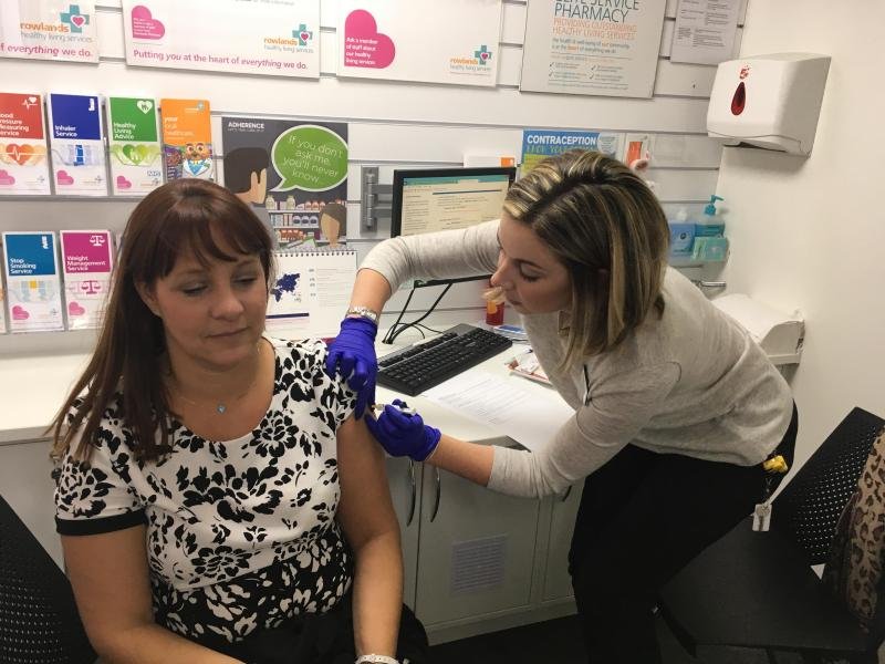 A woman in a patterned dress receives a vaccination from a nurse in a medical office, with informational posters on the wall behind them.
