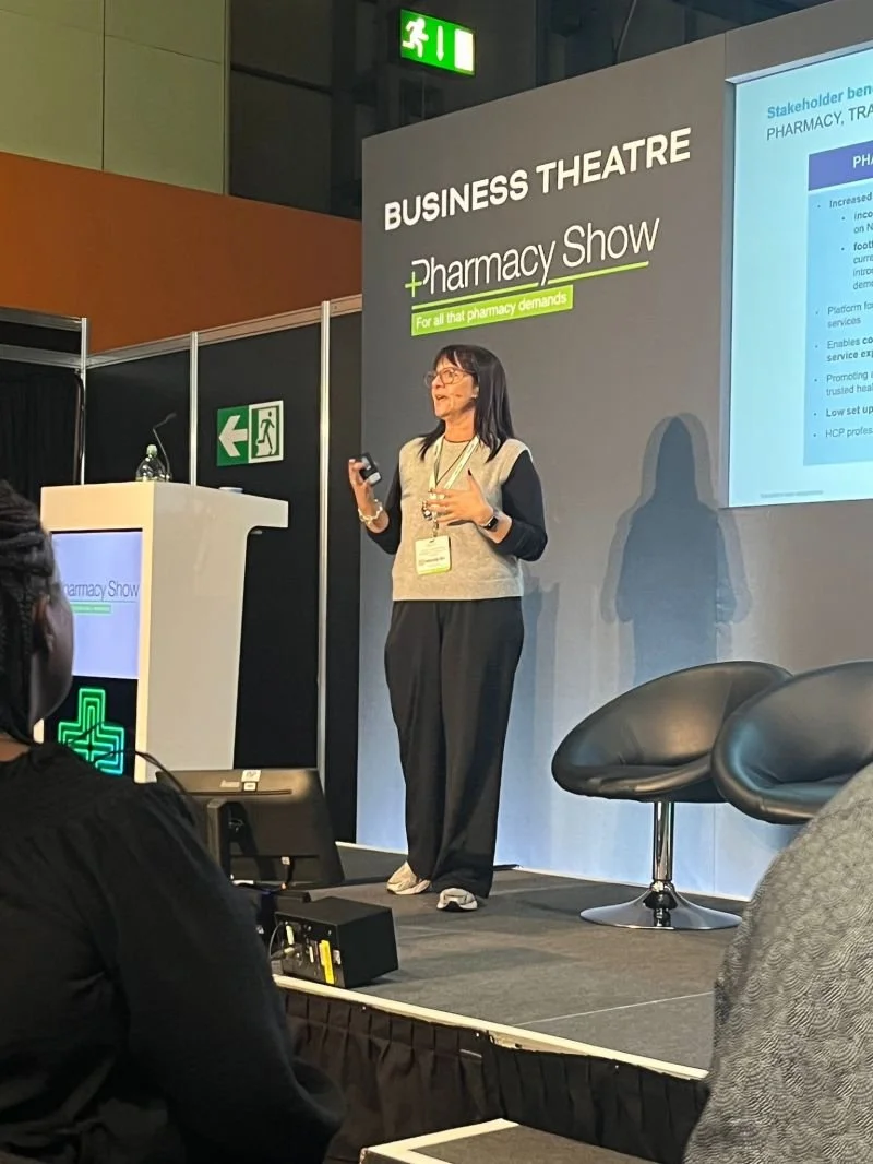 A woman giving a presentation at the Business Theatre during a pharmacy show, with a screen displaying presentation slides behind her.