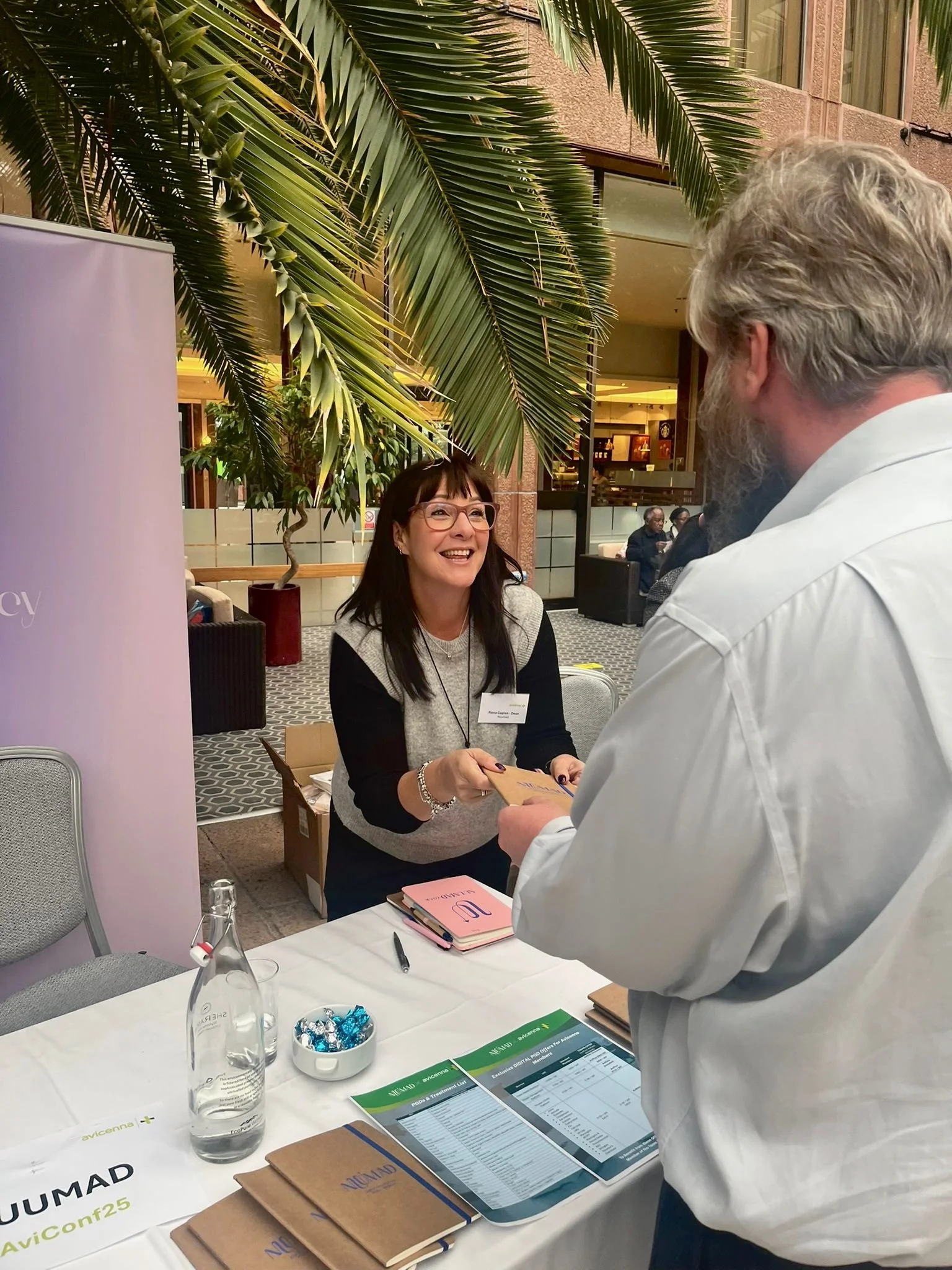 A woman is smiling and handing a certificate or paper to a man. They are at a registration or information table with brochures, a pen, a bowl of wrapped candies, and water bottles. There are large green palm leaves overhead, and other people are visible in the background.