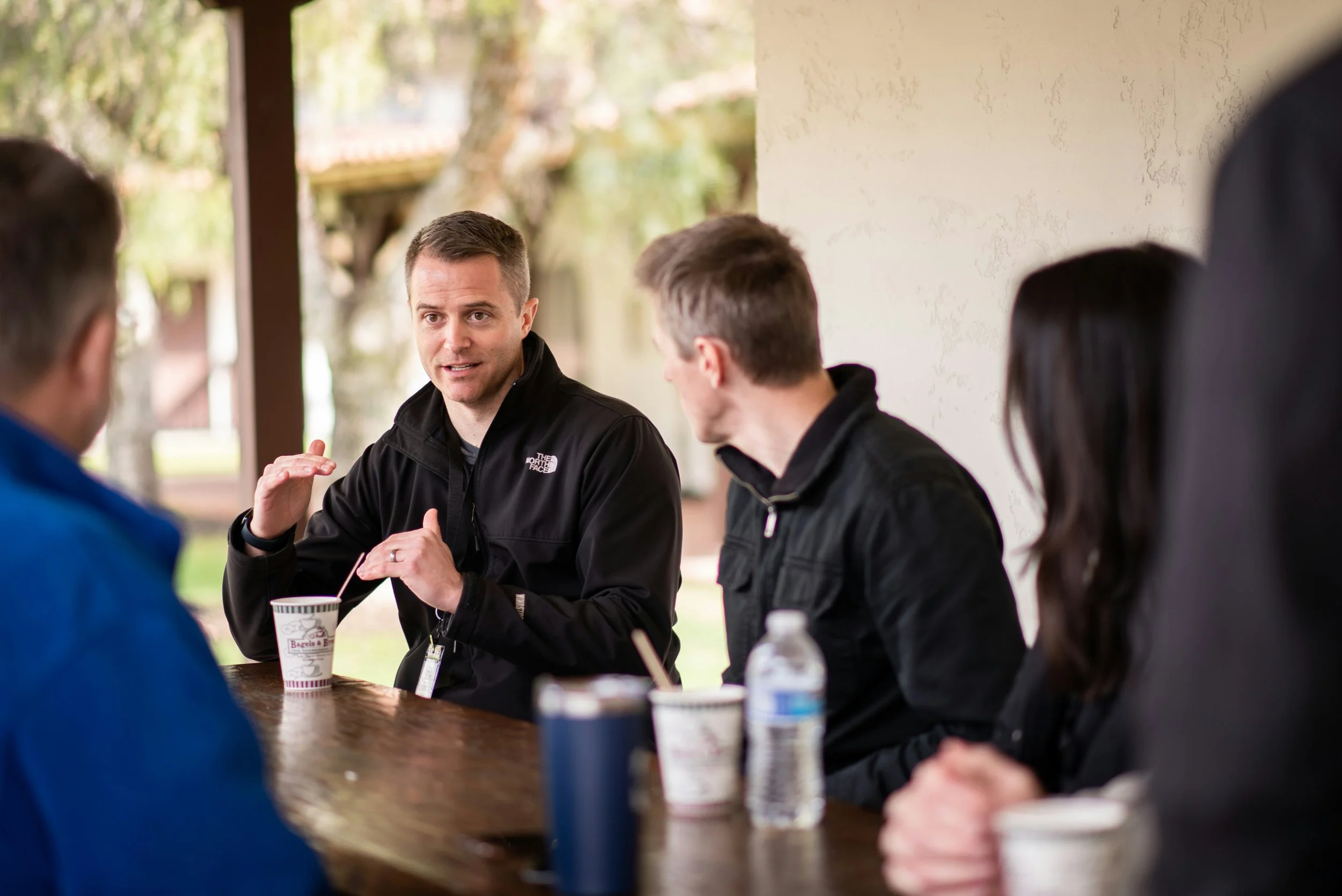 Man in black North Face jacket speaking and gesturing during a discussion with four other people sitting at a wooden table outside with trees in the background.