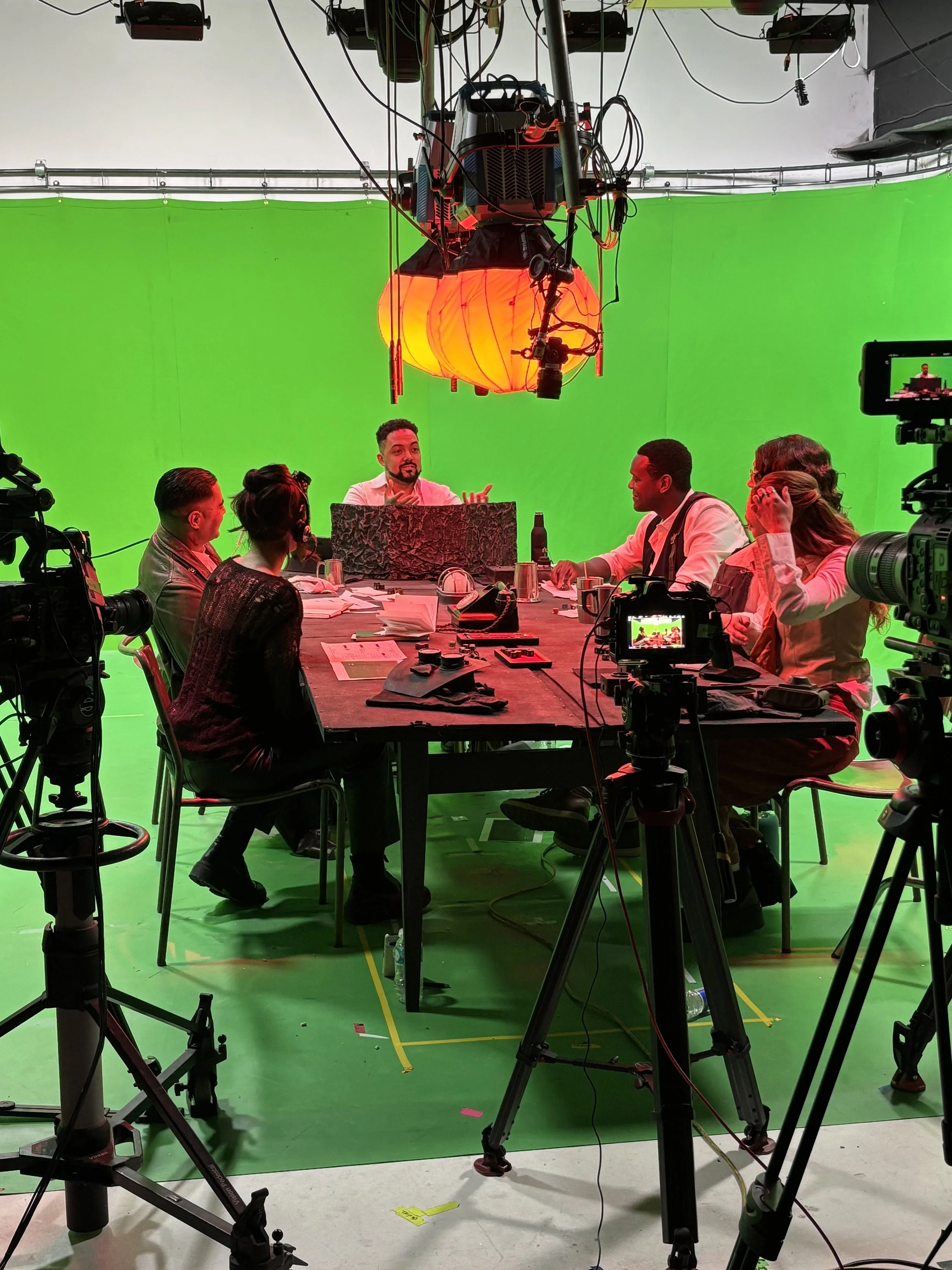 A crew filming video in front of a large green screen, with cameras and lighting equipment set up. Several people are seated at a table, and a man with a beard is presenting.