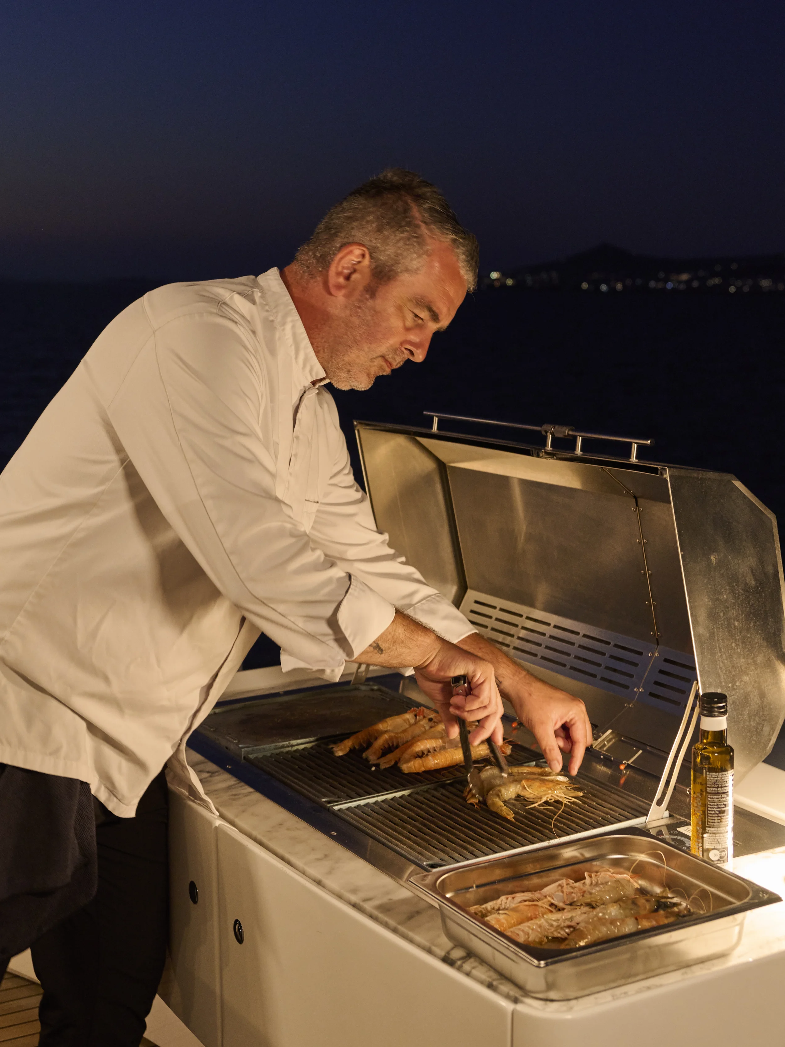 A man in a white shirt grilling shrimp and seafood on a barbecue grill at night, outdoors near water with a distant shoreline.