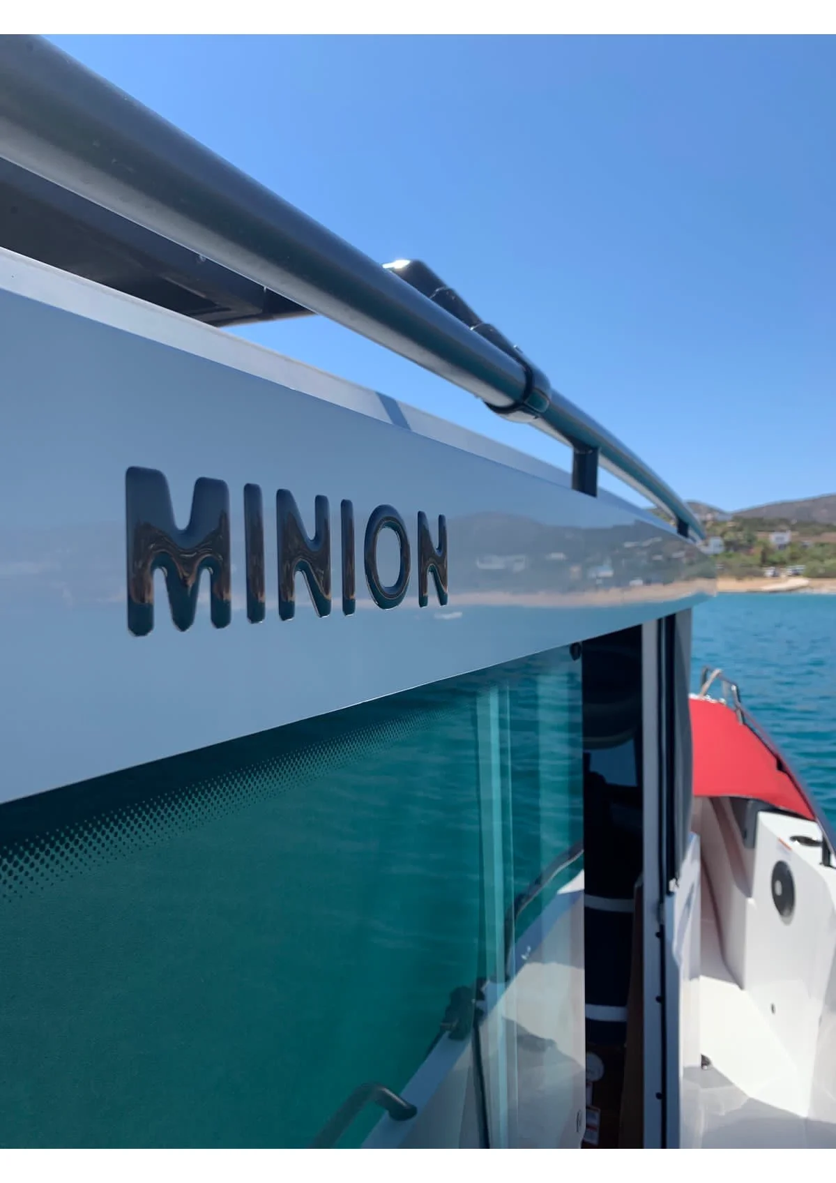 Close-up of a boat with the name "MINION" on the side, docked on clear blue water with a shoreline and hills in the distance under a bright, clear sky.