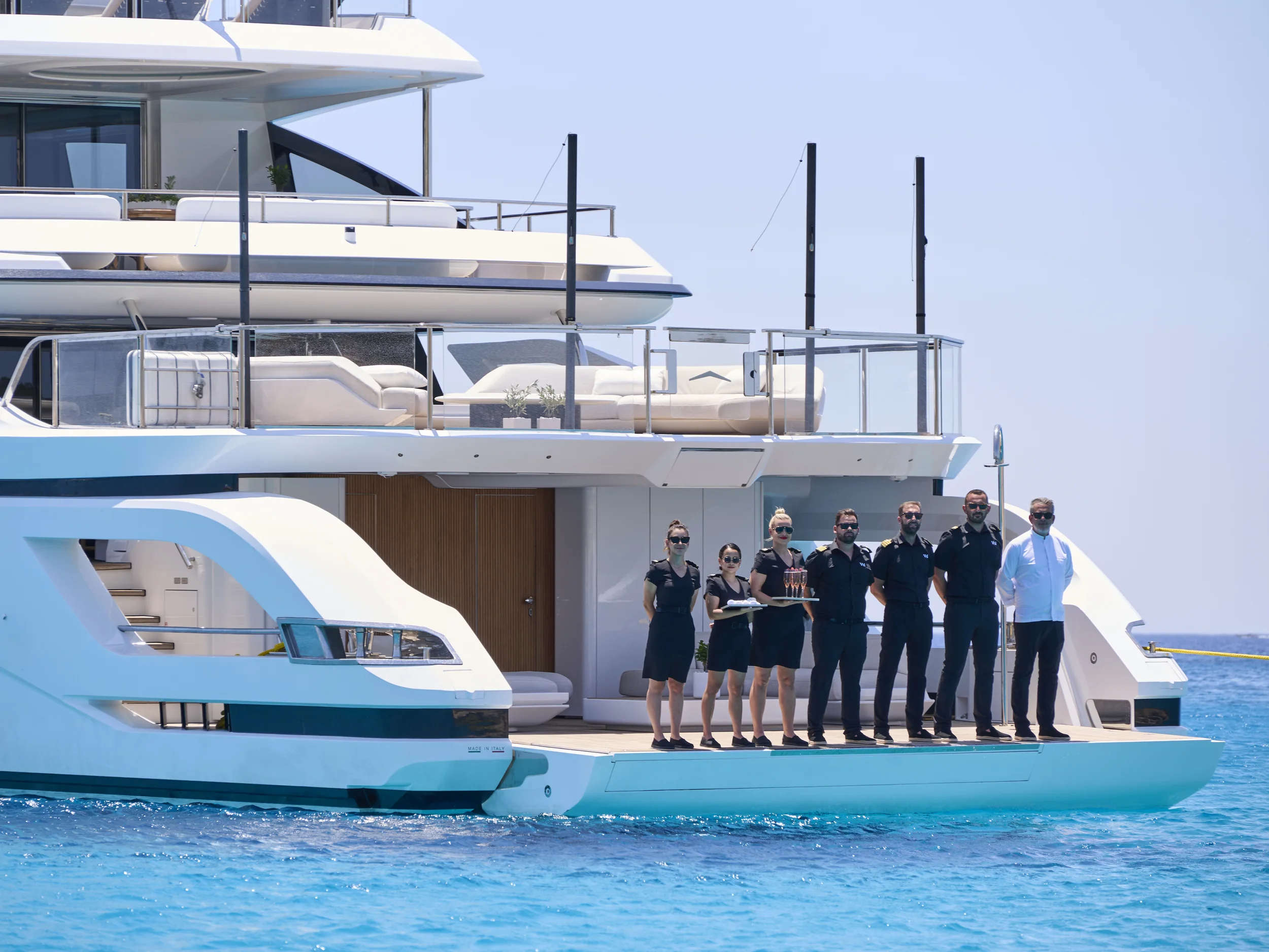 A luxury yacht with crew members standing on the deck, holding a tray with drinks, on calm ocean waters.