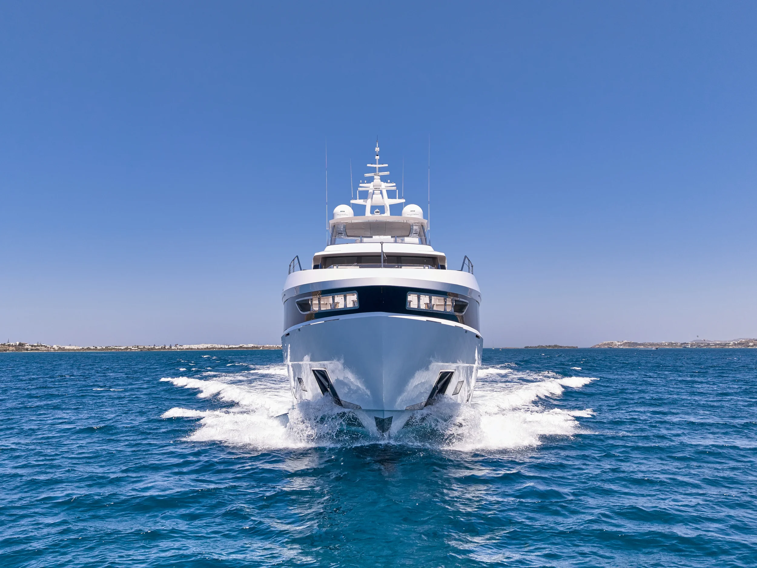 A white yacht sailing towards the camera on clear blue waters with a distant shoreline and blue sky in the background.