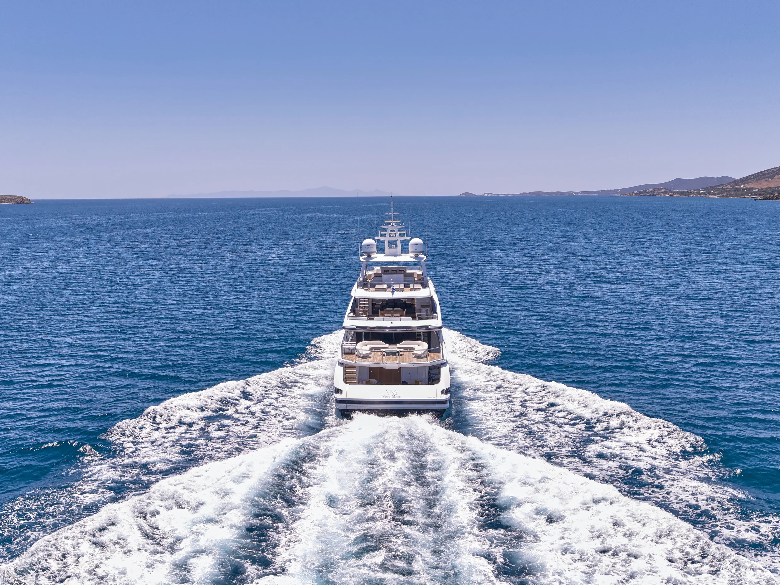 A large luxury yacht sailing away into the open ocean with a distant coastline on the horizon under a clear blue sky.