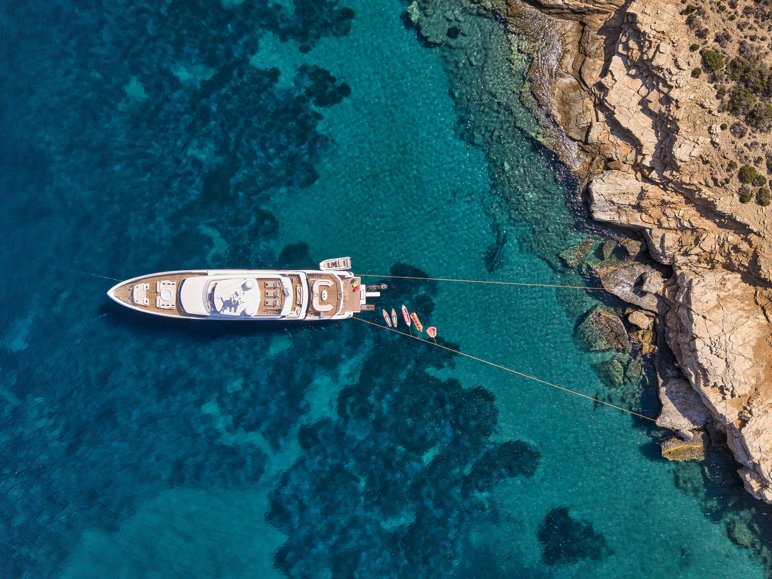 Aerial view of a yacht anchored near a rocky coastline with clear blue water.